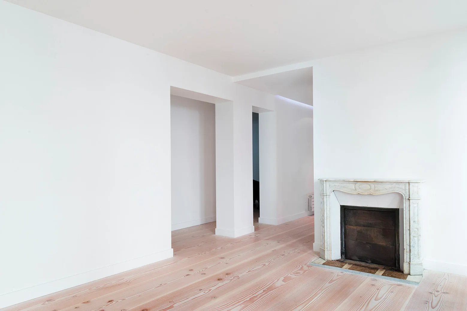 A minimalist white room featuring wide-plank light wood flooring, casing-free doorways, and a traditional carved stone fireplace.