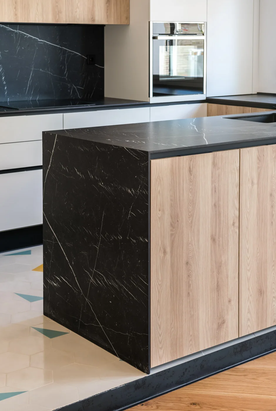 A modern kitchen featuring a black marble-patterned waterfall island with light wood cabinet doors, set against hexagonal patterned floor tiles.