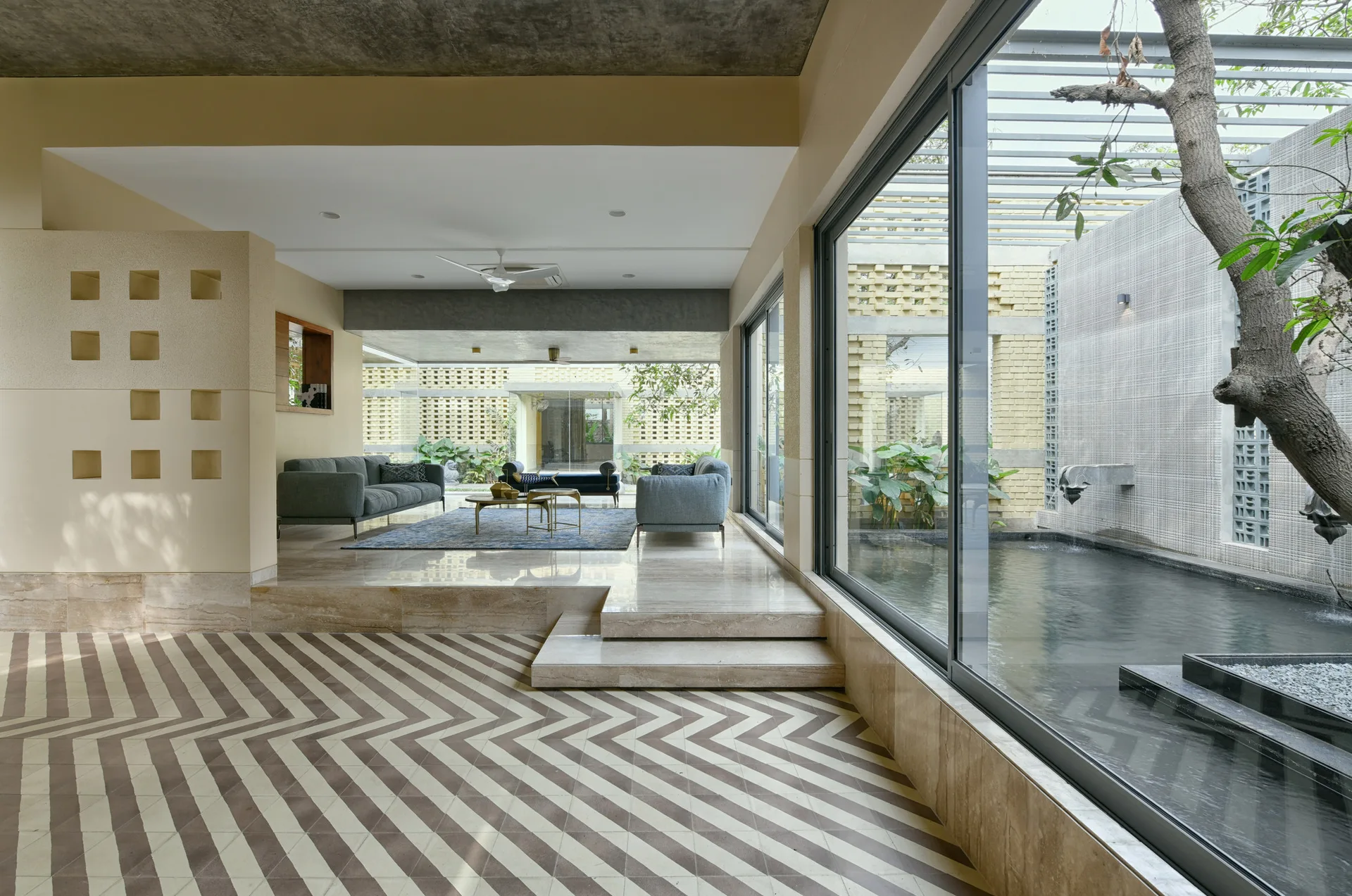 A raised modern living area with gray sofas overlooks an outdoor water feature and tree through large glass windows, beside a chevron-patterned floor.
