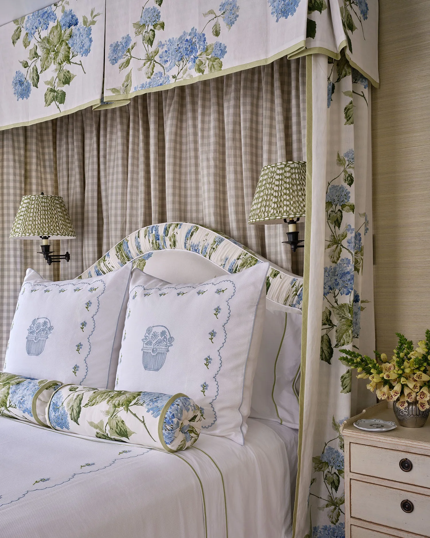 A canopy bed styled with blue hydrangea print bedding and an arched headboard, set against a neutral gingham fabric backdrop and matching wall sconces.