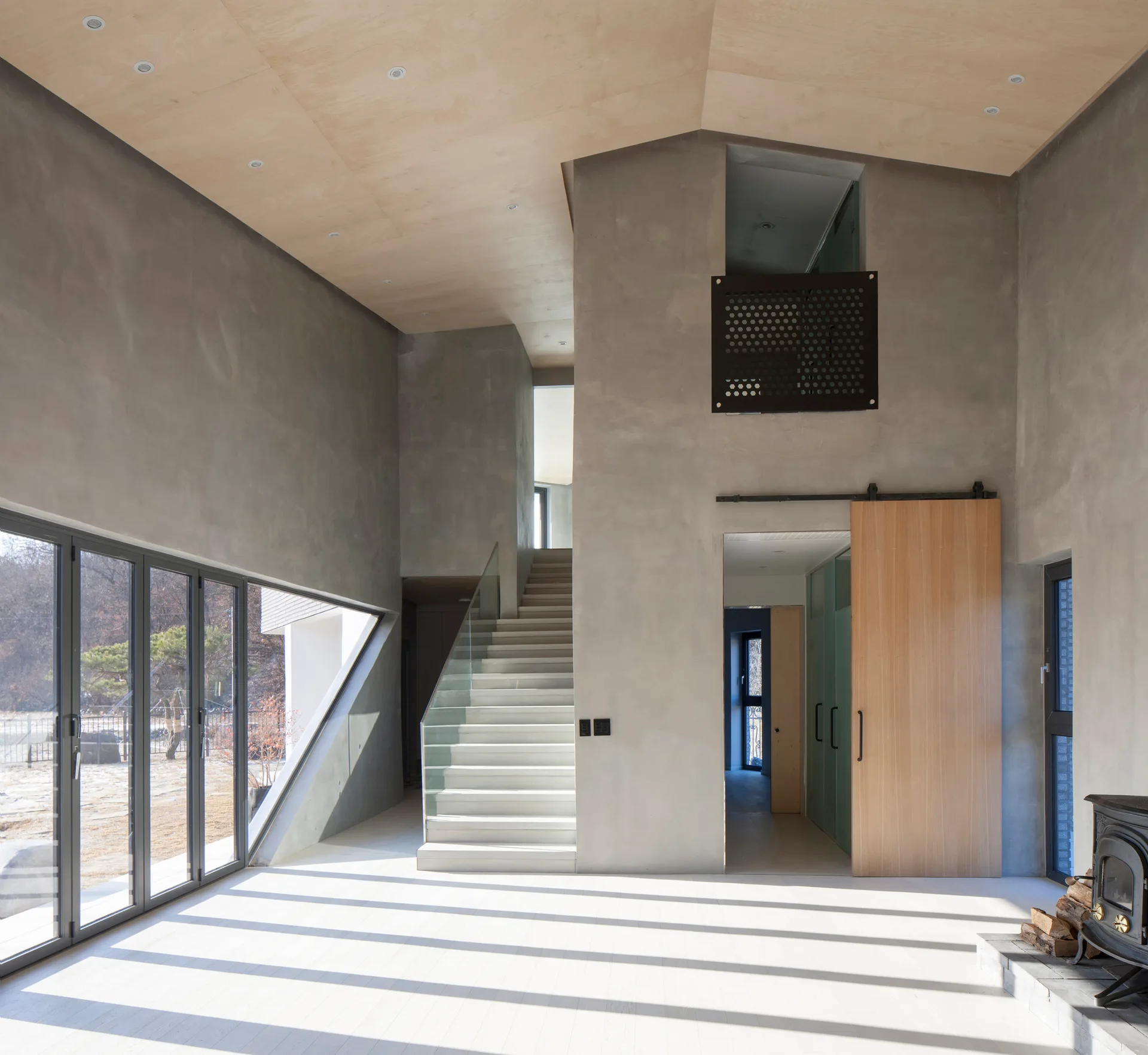 A minimalist double-height room features textured grey concrete walls, a sloped wood ceiling, and a central staircase with a glass railing.