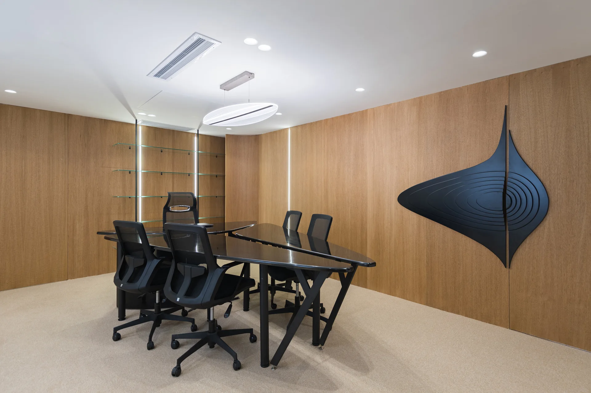 A minimalist meeting room with wood-paneled walls, black modular conference tables, mesh office chairs, and an abstract black wall sculpture.