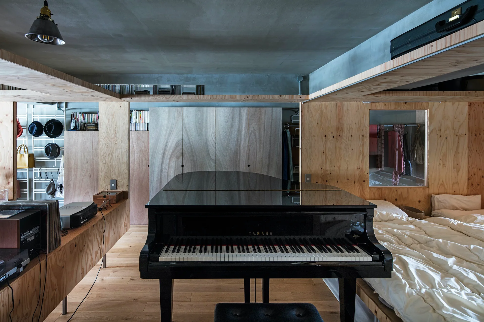 A compact bedroom features a black grand piano in the center, surrounded by light wood plywood wall panels, built-in shelving, and a low platform bed.