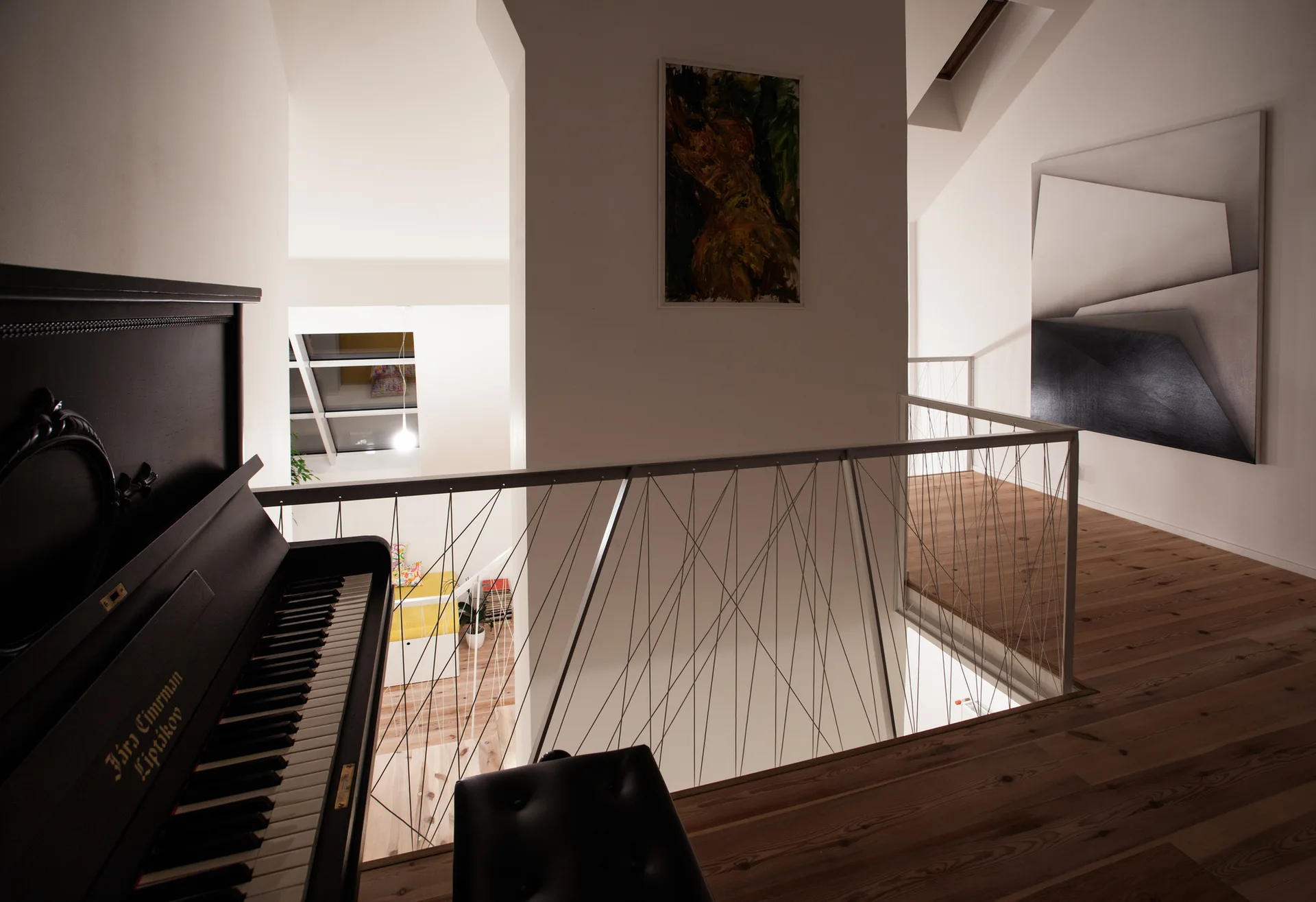 A contemporary loft landing features a black upright piano, light wood plank floors, and a white metal railing with geometric tension wires.