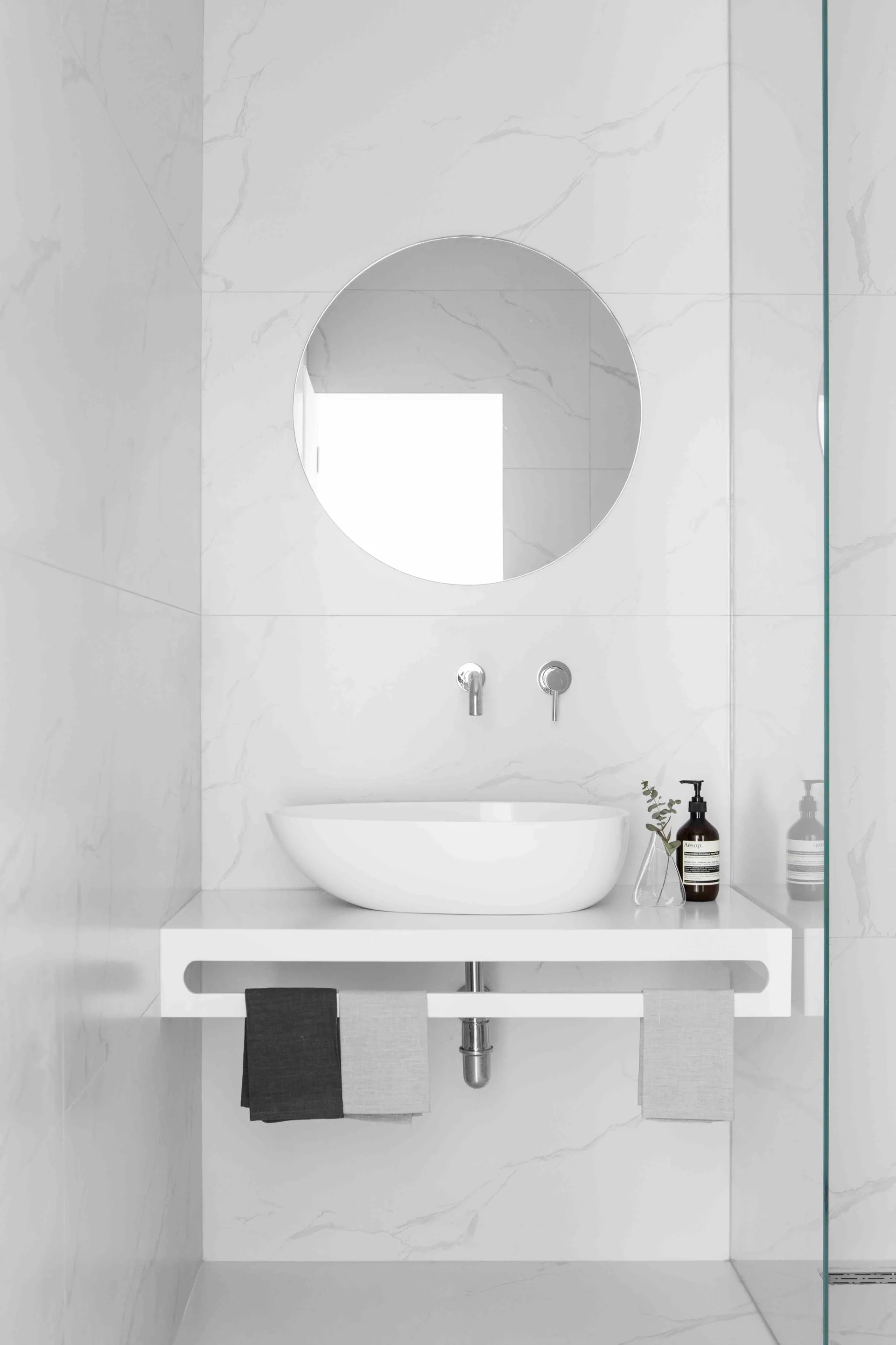 A minimalist bathroom nook features a floating white vanity with an oval vessel sink, a circular wall mirror, and grey marble-patterned wall tiles.