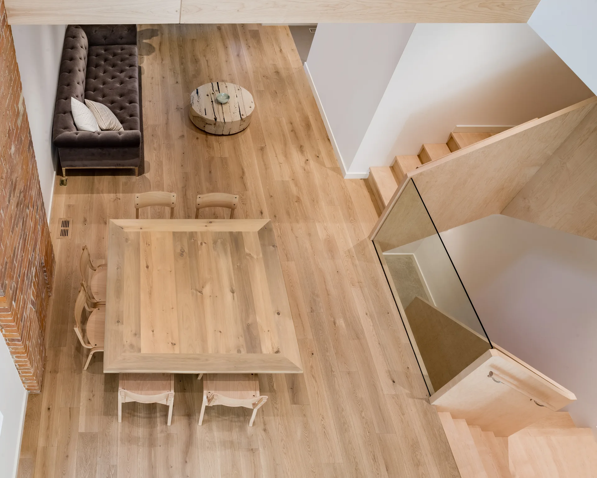 An overhead view of an open living and dining area featuring light hardwood floors, a wooden dining table, a tufted grey sofa, and a staircase.