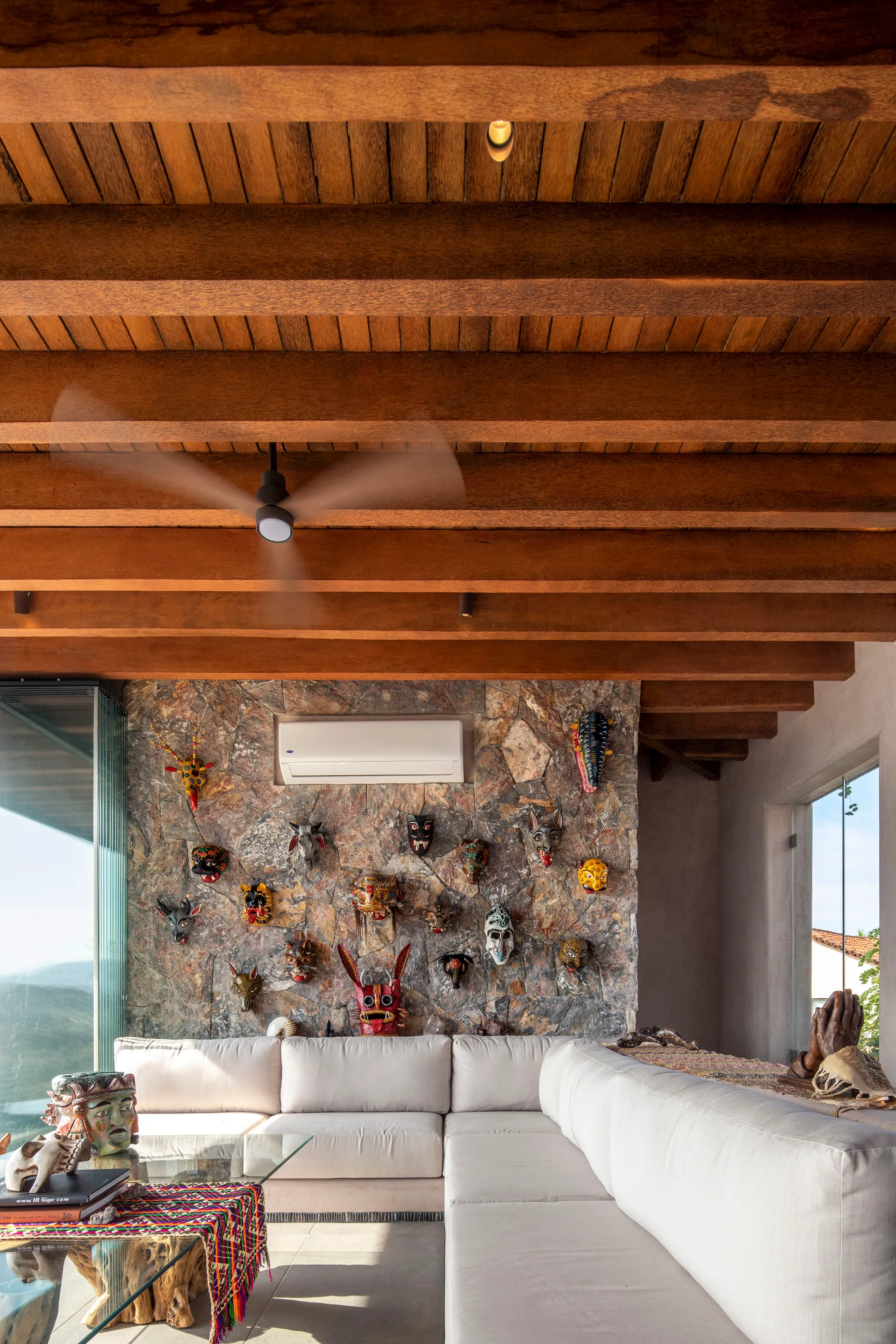 An eclectic living room with a cream sectional sofa, a textured stone wall decorated with masks, and an exposed timber beam ceiling.