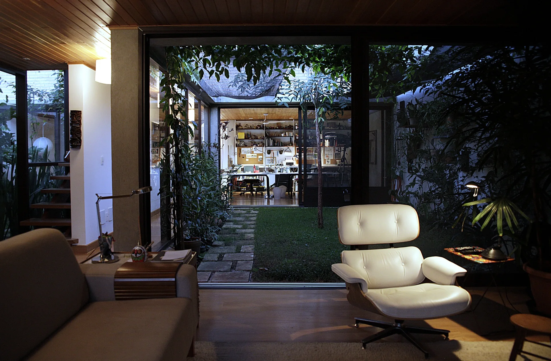 A mid-century modern living room featuring a white leather lounge chair and glass doors that open to a lush courtyard with stone pavers.