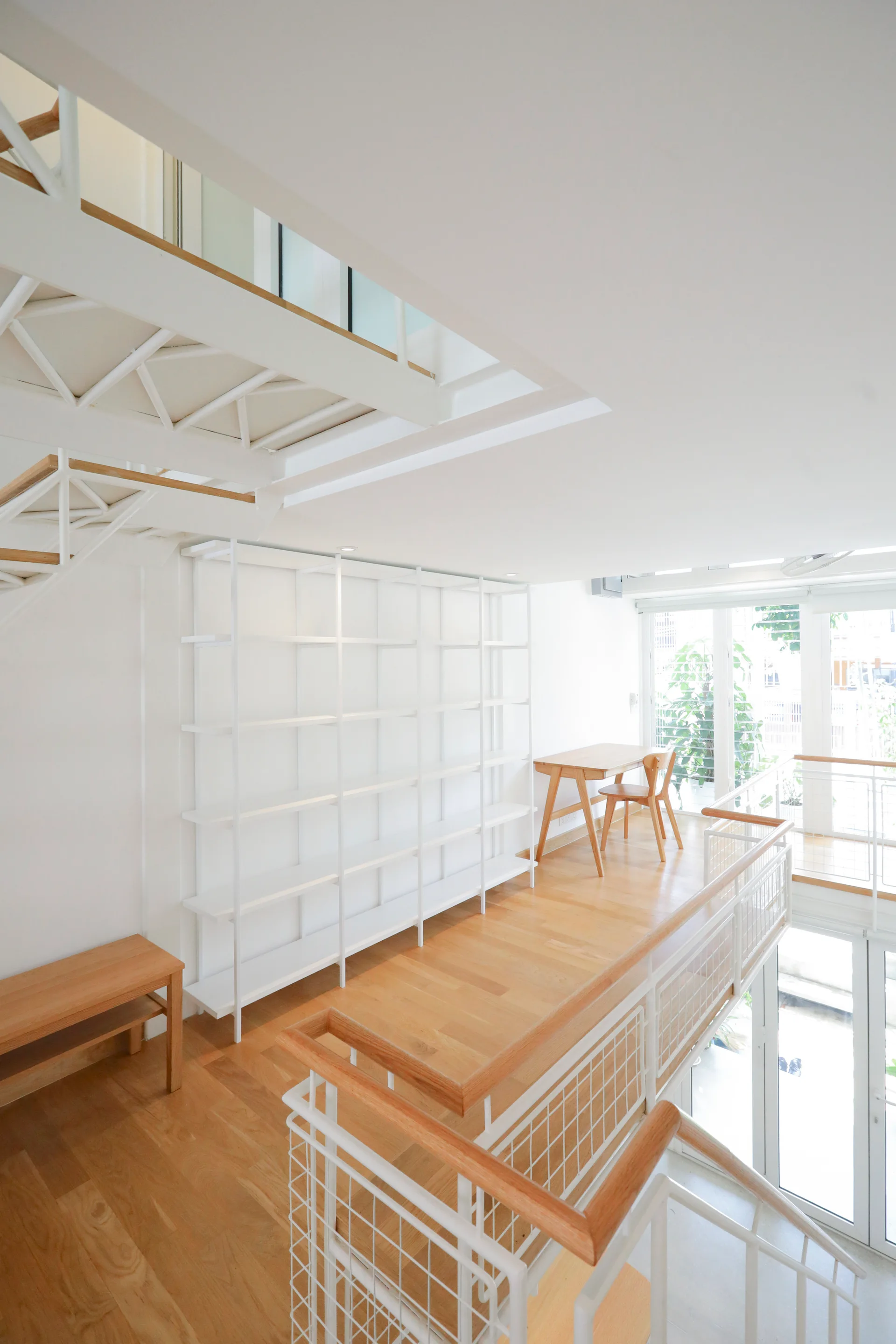 A minimalist loft landing features light wood flooring, a tall white open shelving unit, a small wood desk, and a metal grid railing.