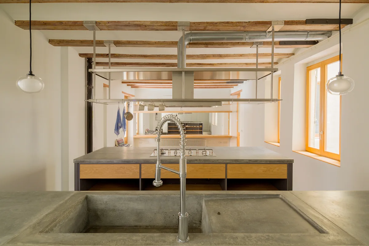 A modern rustic kitchen featuring a concrete island with an integrated sink, an industrial faucet, and exposed wooden ceiling beams.