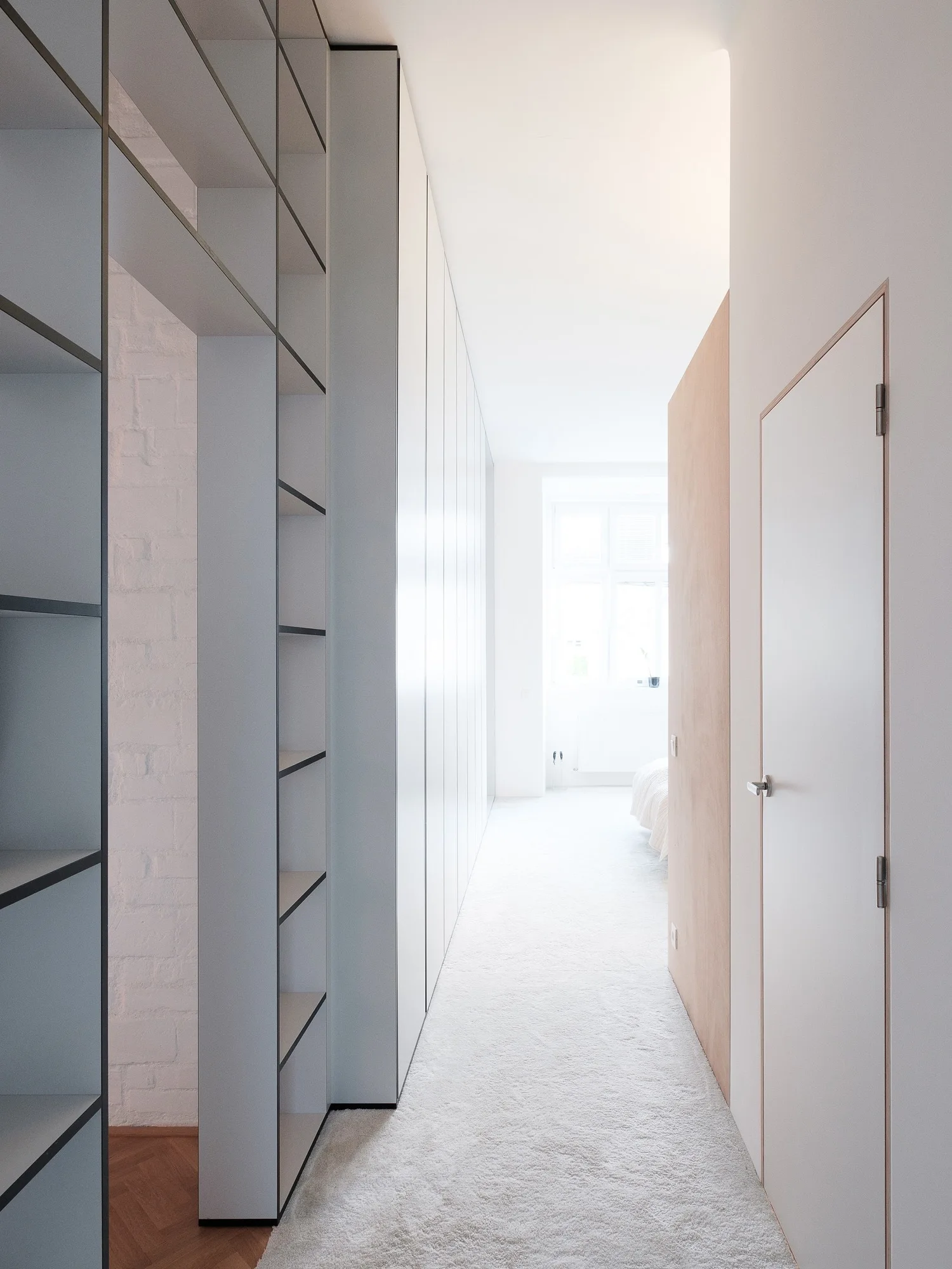 A bright white hallway with built-in storage cabinets on one side, a light wood door on the other, and carpet leading to a bedroom window.