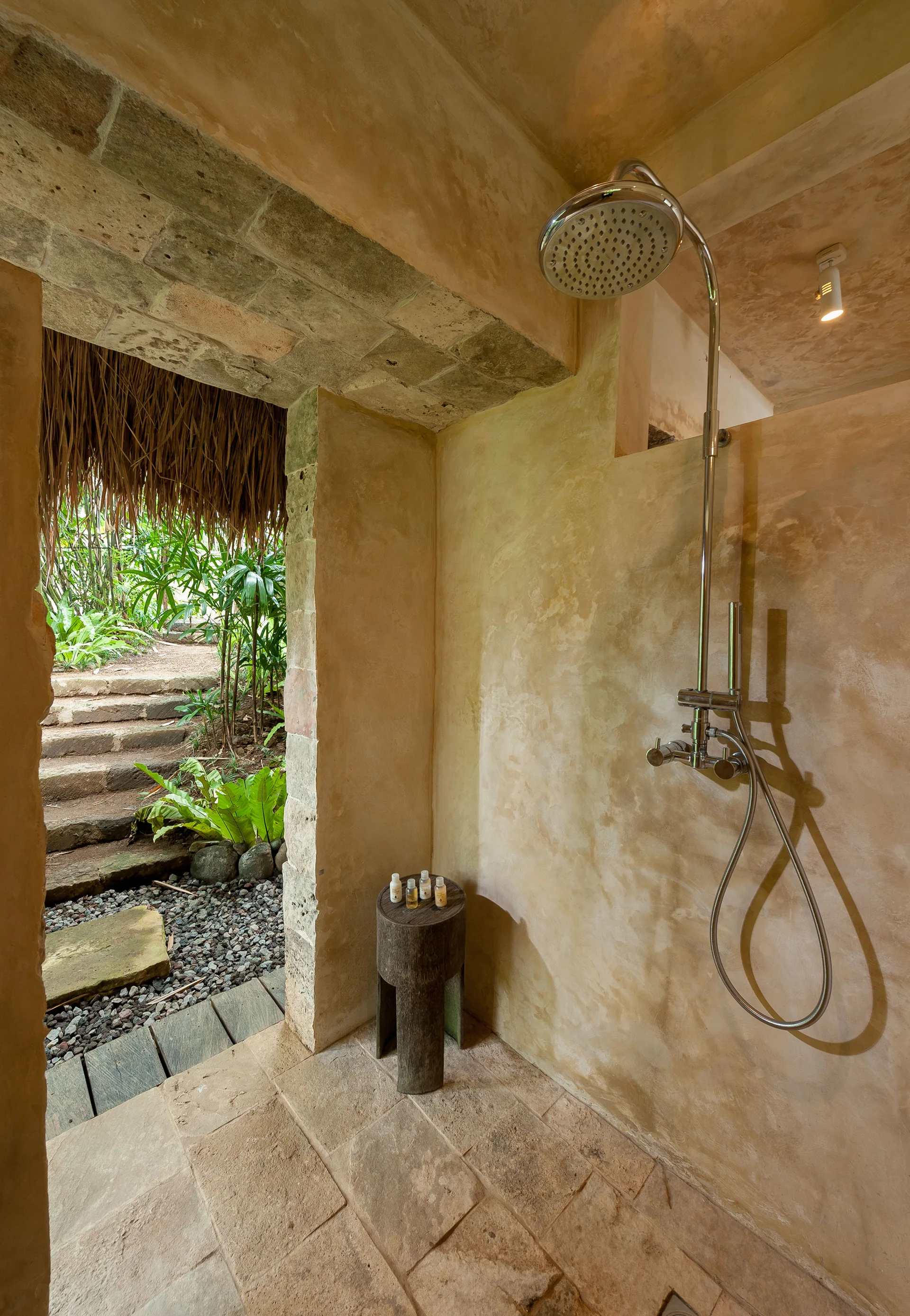 An open-air shower featuring beige textured plaster walls, a chrome shower fixture, a wooden stool, and a view leading to a lush green garden path.