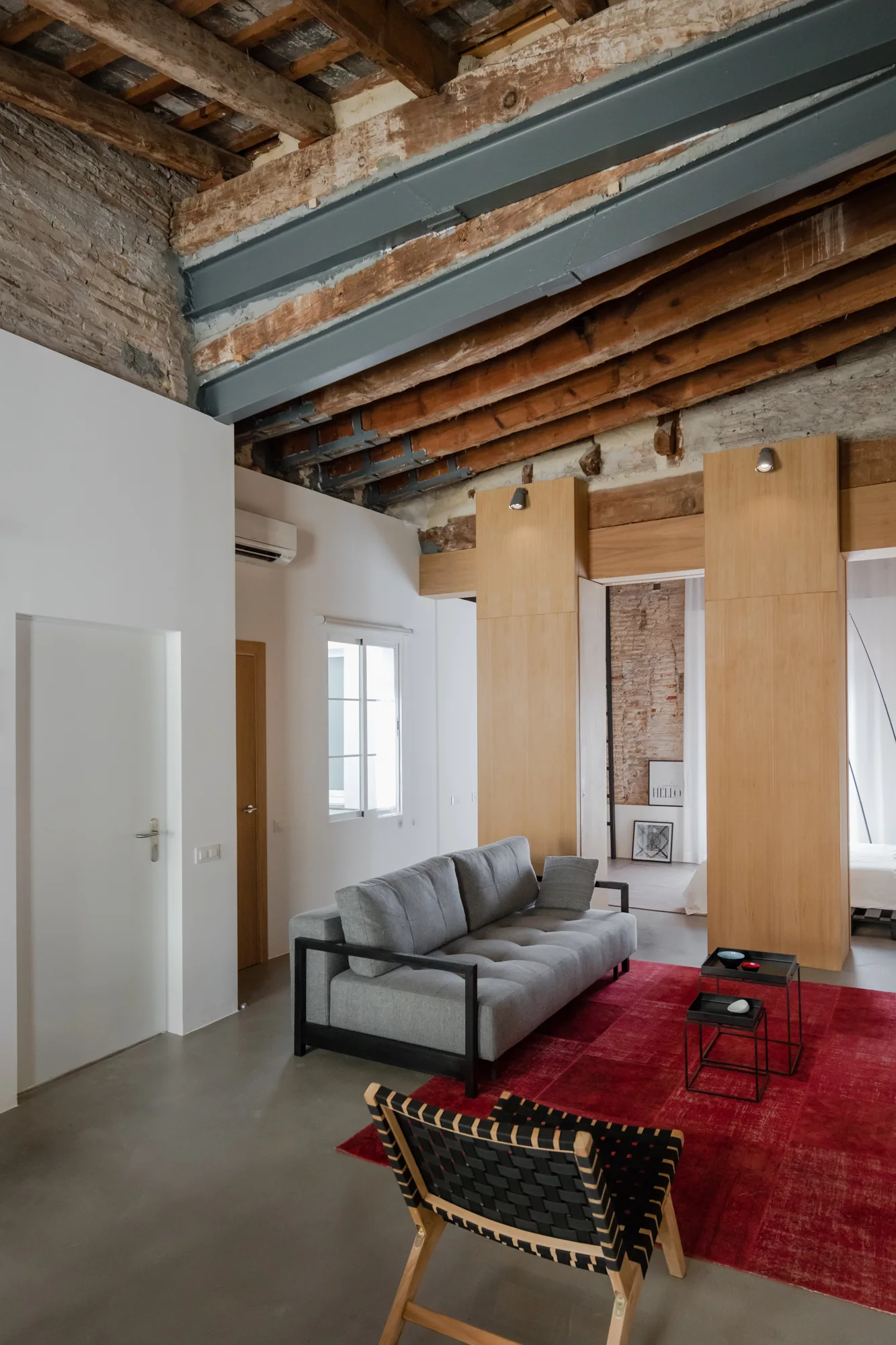 A minimalist grey sofa and woven accent chair sit on a large red rug beneath a high ceiling with exposed wooden beams.