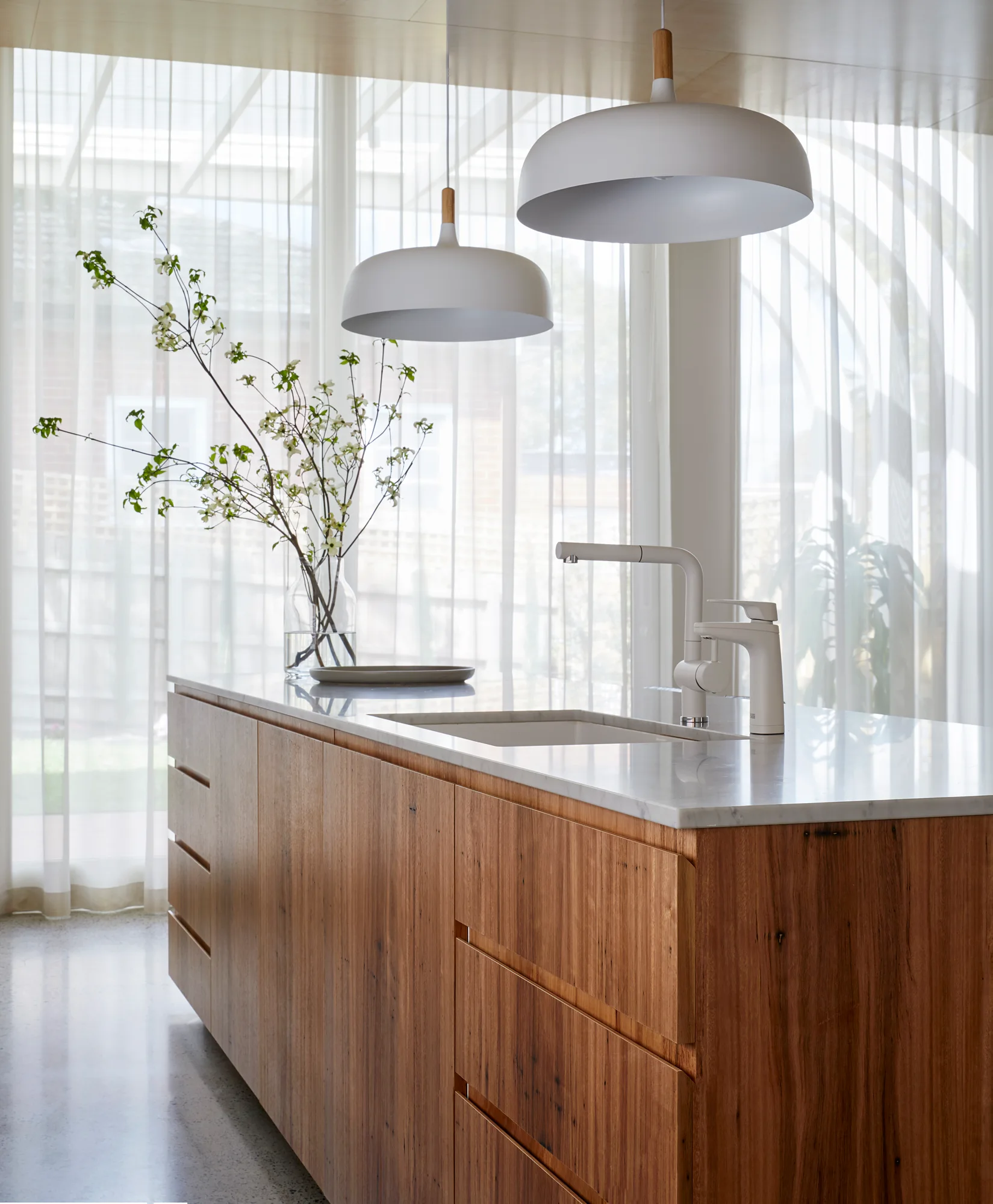 A natural wood kitchen island with a white stone countertop, integrated sink, white faucet, and matching pendant lights sits before sheer white curtains.