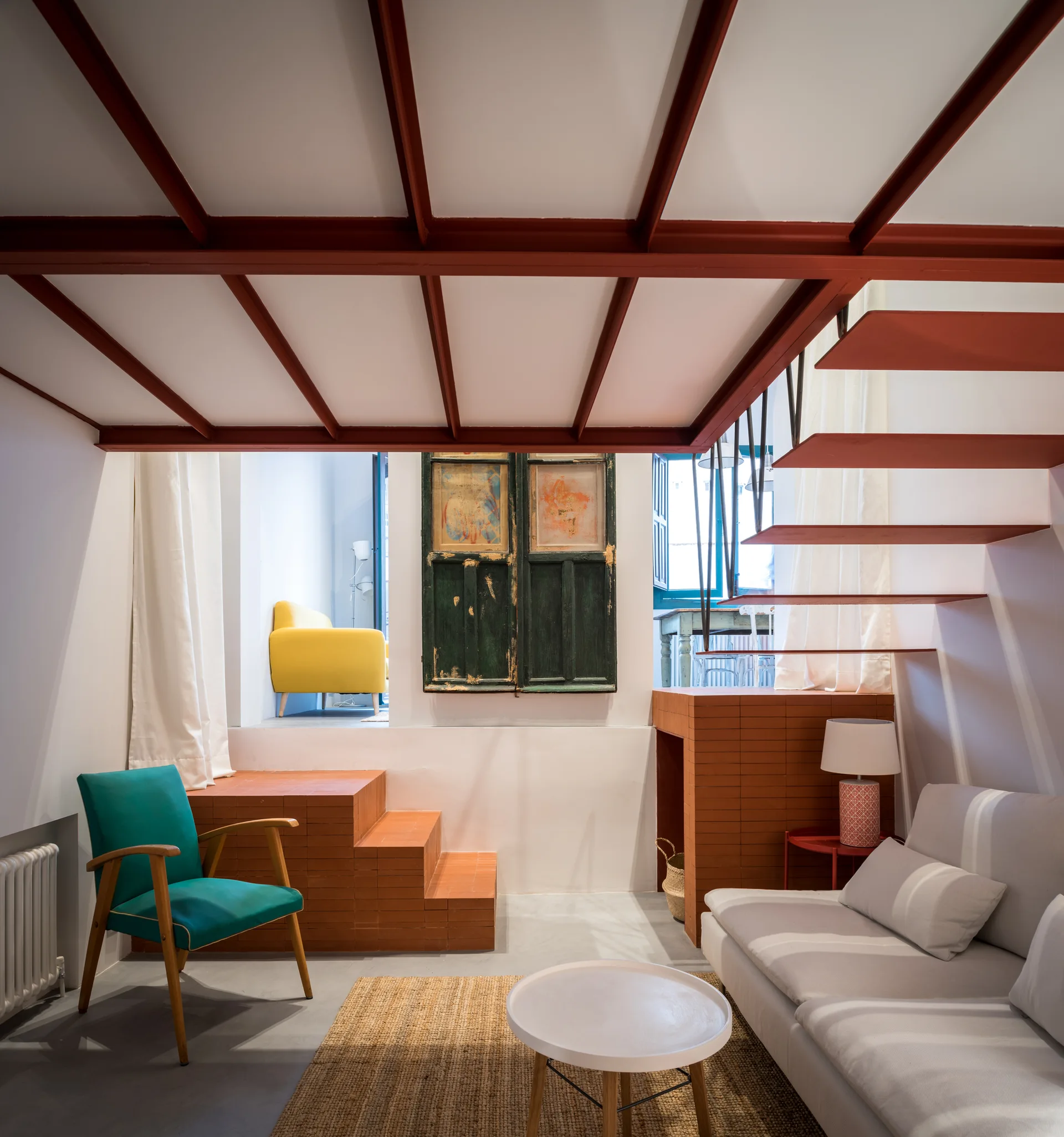 An eclectic loft living room features a white sofa, a teal armchair, and a red metal mezzanine structure with stepped brick platforms.