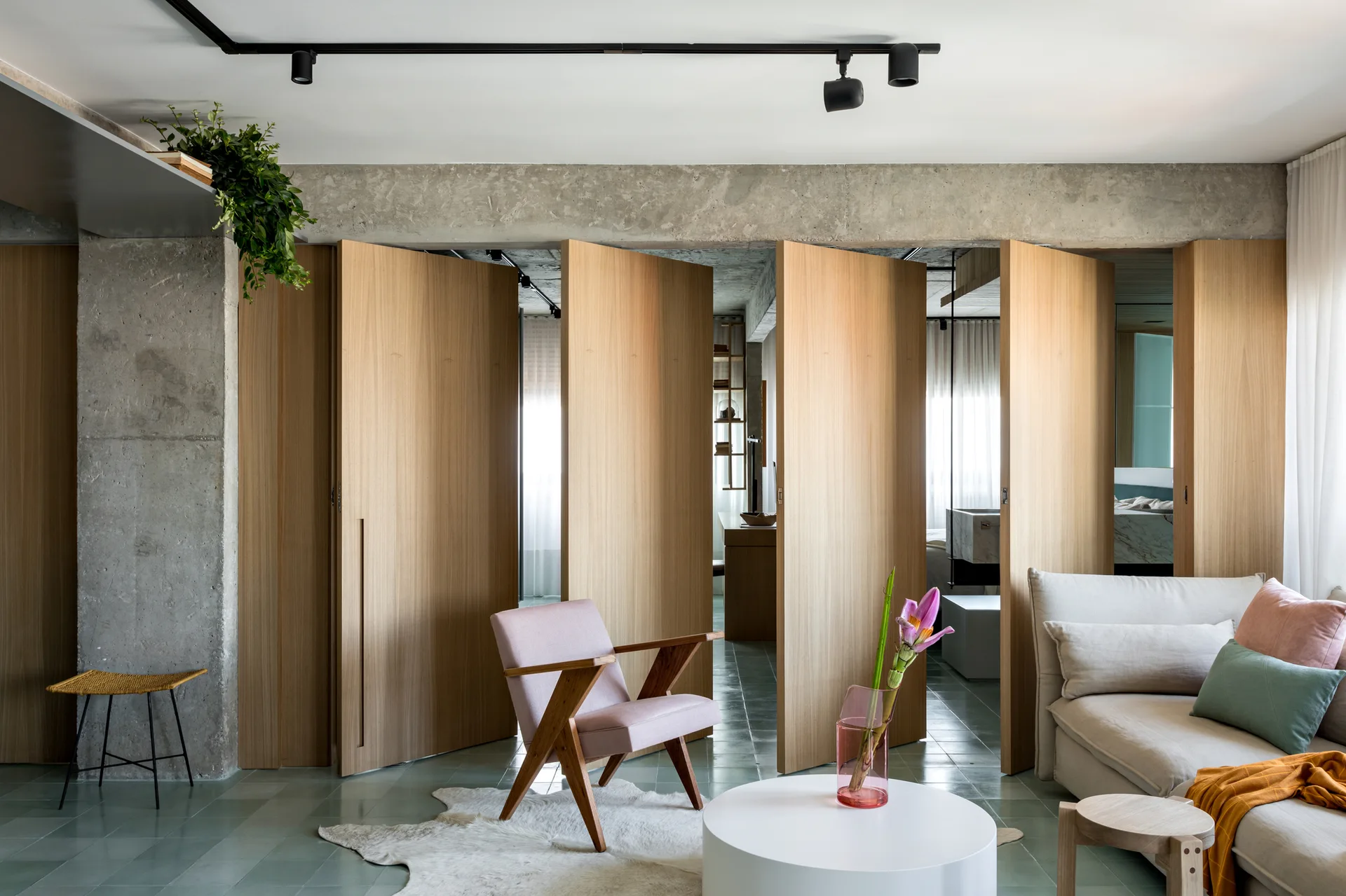 A modern living room features a folding wood panel room divider, an exposed concrete beam, a pink armchair, and green floor tiles.