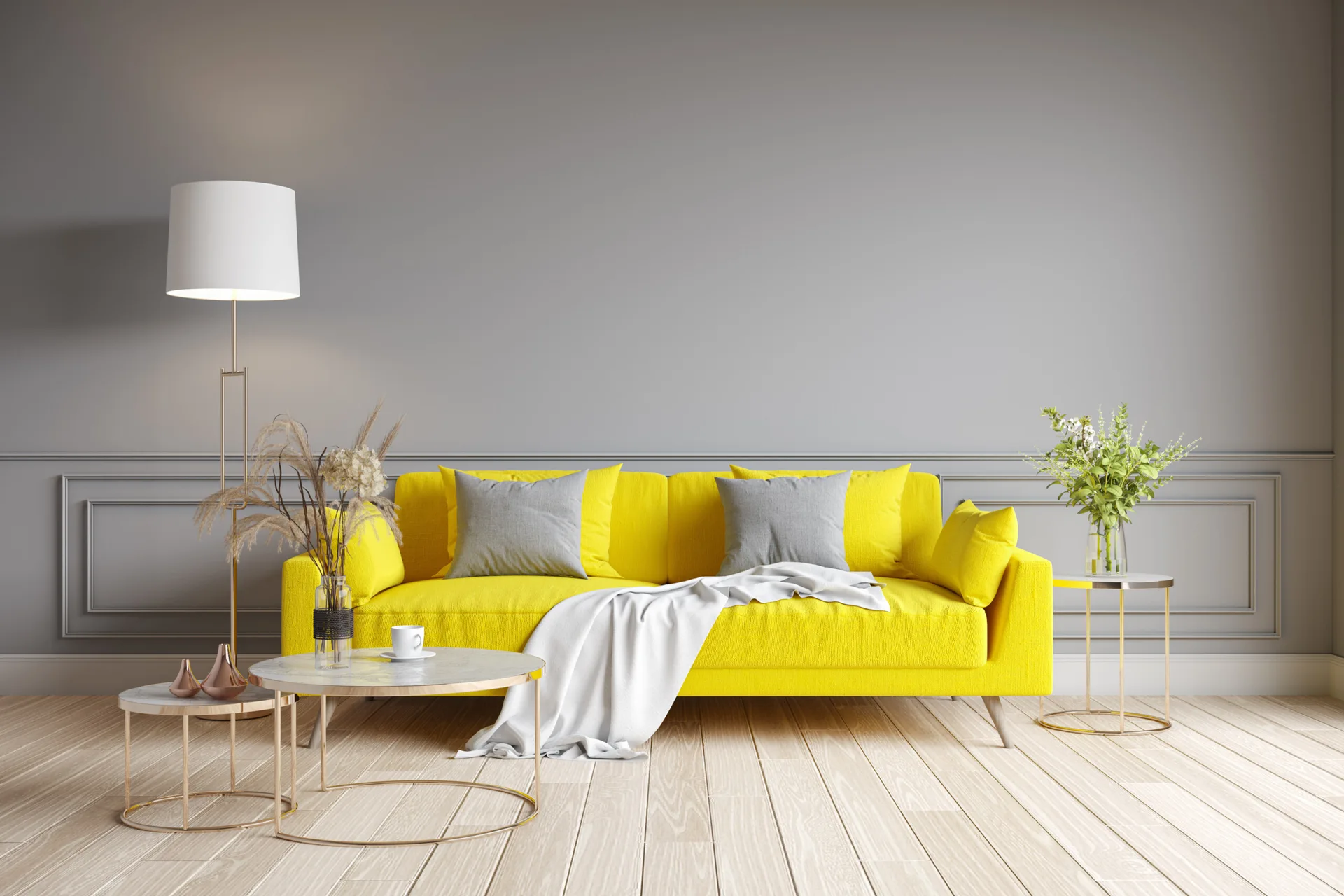 A bright yellow sofa with gray pillows sits against a paneled gray wall in a room with light wood floors and gold-framed coffee tables.
