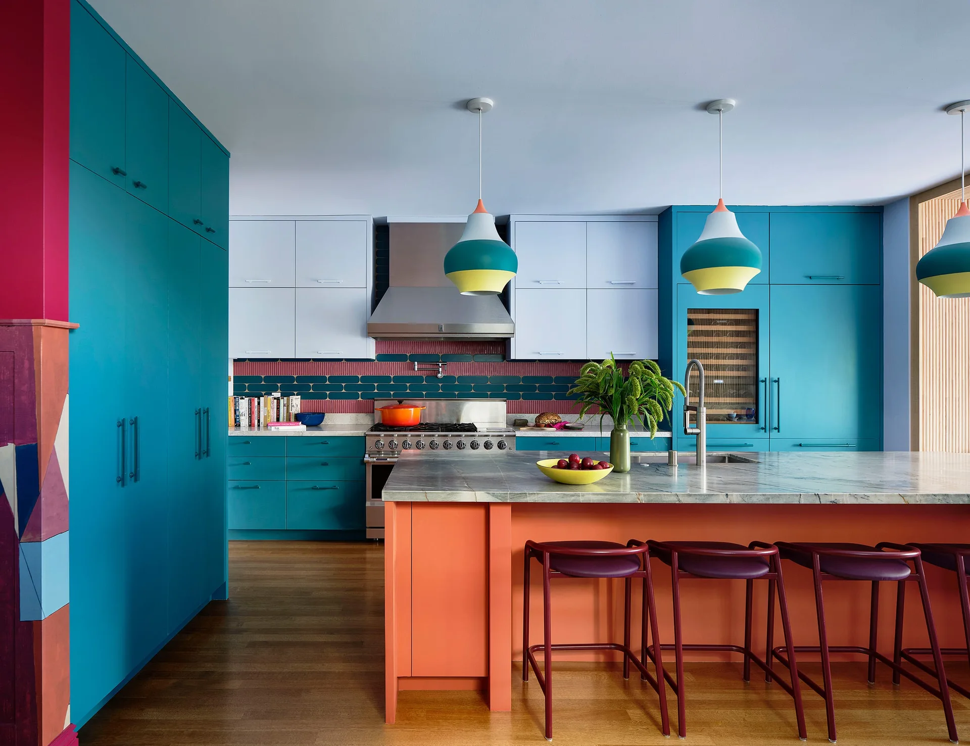 A colorful modern kitchen features matte teal cabinets, a bright orange kitchen island with a grey stone countertop, and three hanging pendant lights.