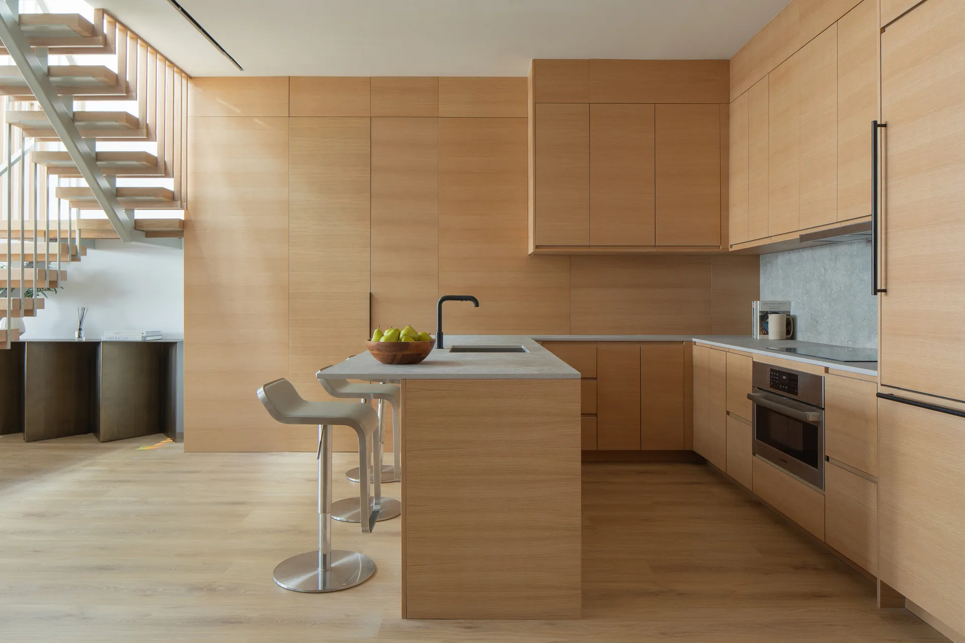 A minimalist kitchen features seamless light wood cabinetry, a central island with gray stone countertops, and an open riser staircase in the background.
