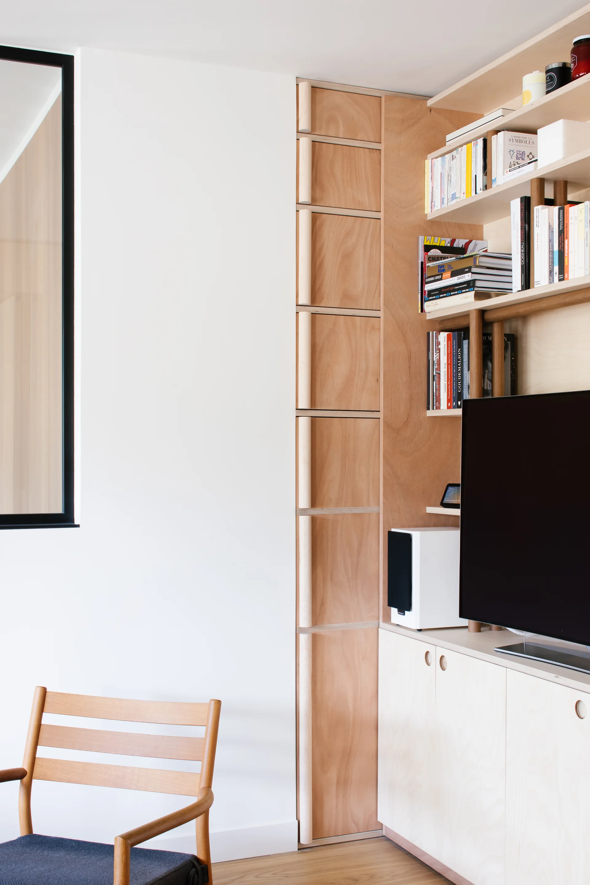A modern corner featuring custom light-toned plywood cabinetry with open shelves, integrated wood pull handles, a television, and a wooden chair.