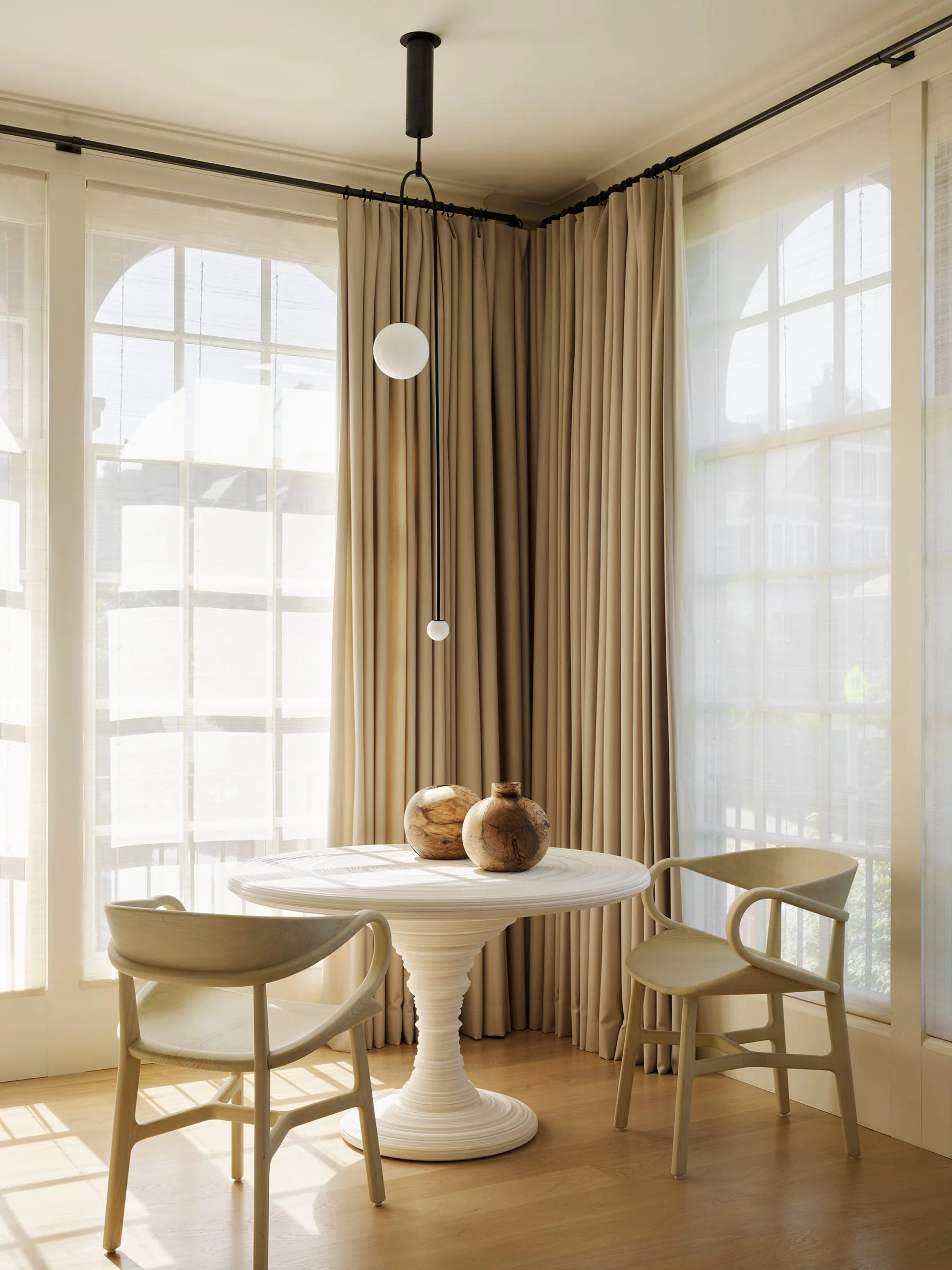 A bright dining corner features a white ribbed pedestal table, two curved wooden chairs, and floor-to-ceiling beige curtains next to arched windows.