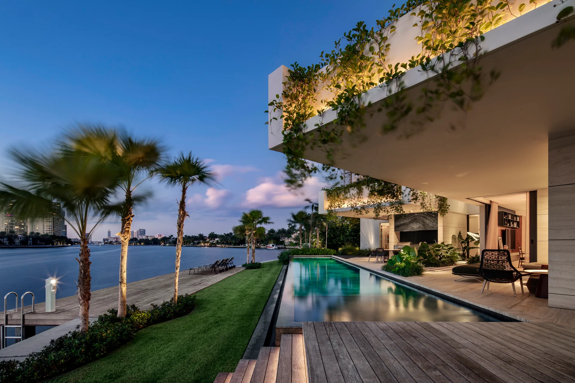 A modern waterfront home at dusk features a long infinity pool, a wooden deck, palm trees, and a cantilevered concrete structure with climbing vines.