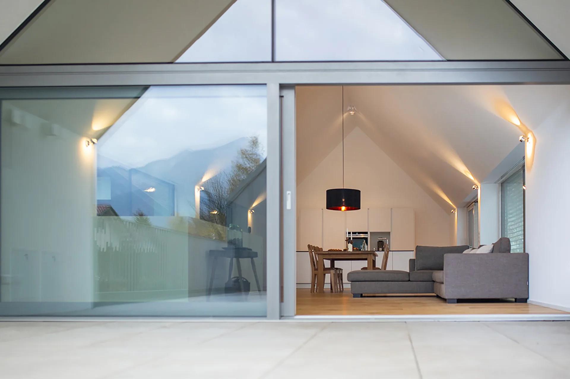 A view through large sliding glass doors into a modern living and dining room featuring a vaulted ceiling, gray sectional sofa, and wooden table.