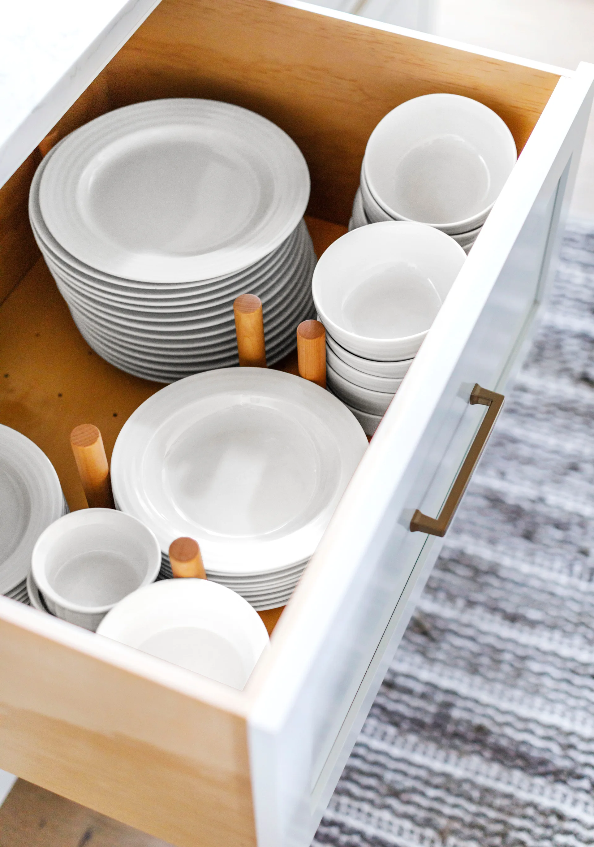 An open white kitchen drawer displays stacks of white plates and bowls separated by vertical wooden peg dividers.