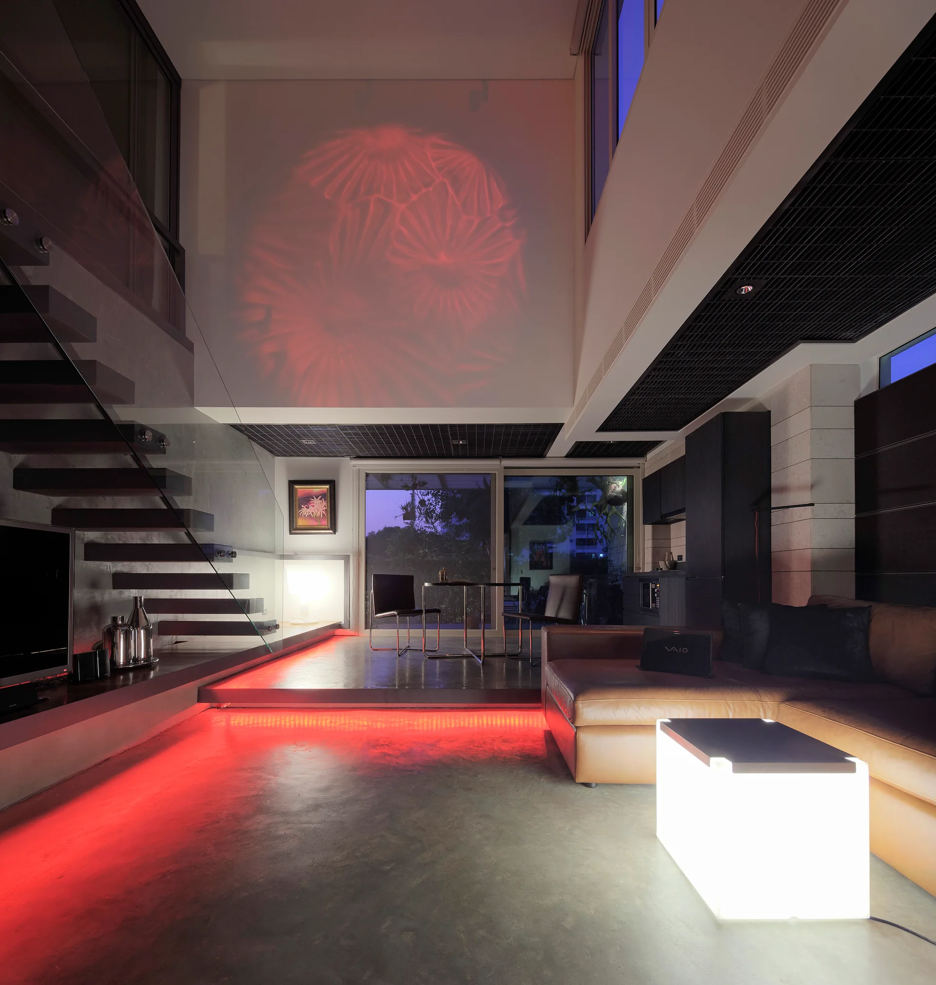 A modern multi-level living room features a floating staircase with glass railing, a tan leather sofa, and glowing red LED floor lighting.