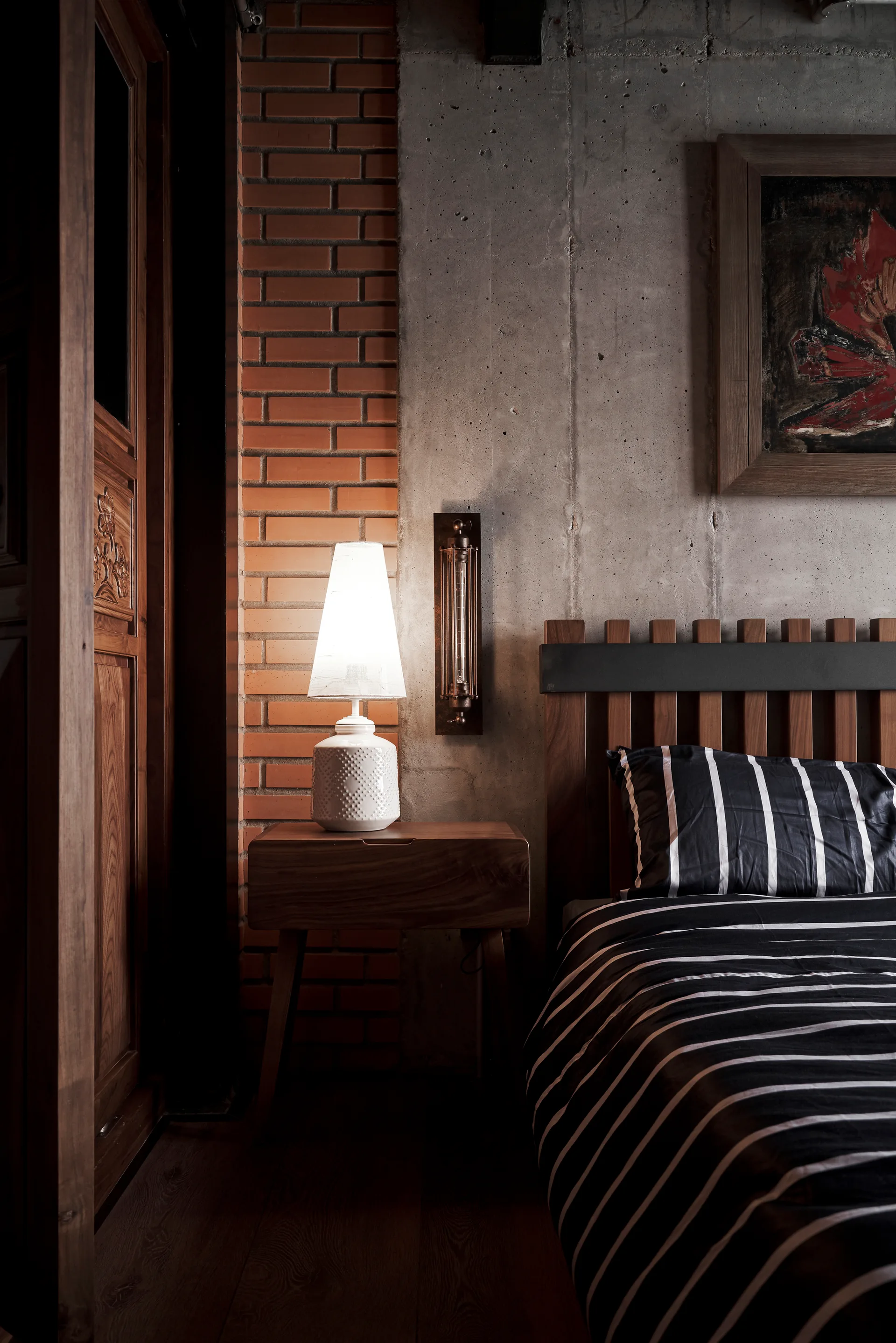 An industrial bedroom features a wood slatted bed next to a nightstand with a white ceramic lamp against exposed brick and raw concrete walls.
