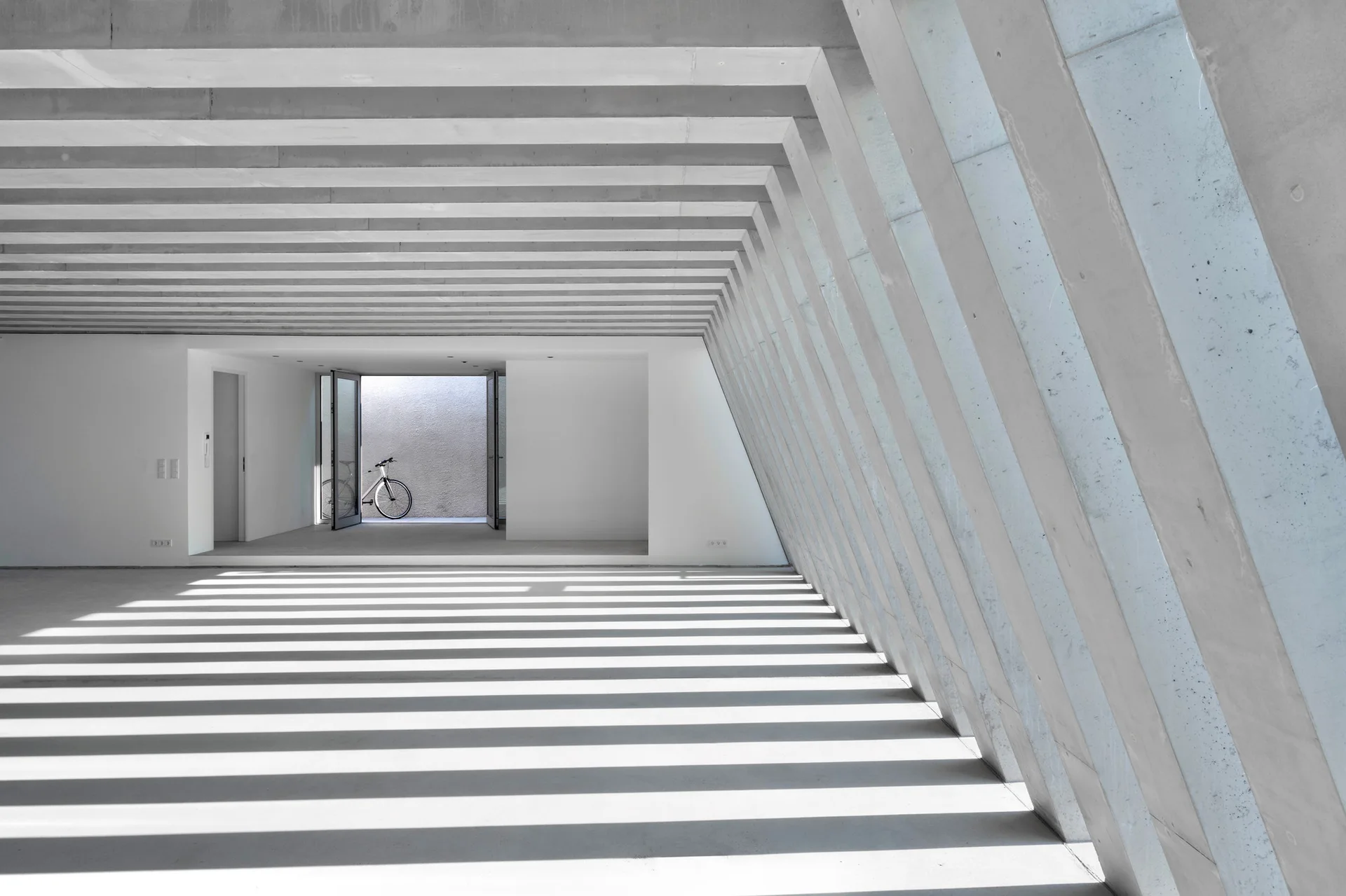 Parallel concrete beams and angled columns cast a striped pattern of shadows across the floor of an empty minimalist room with a bicycle in the background.