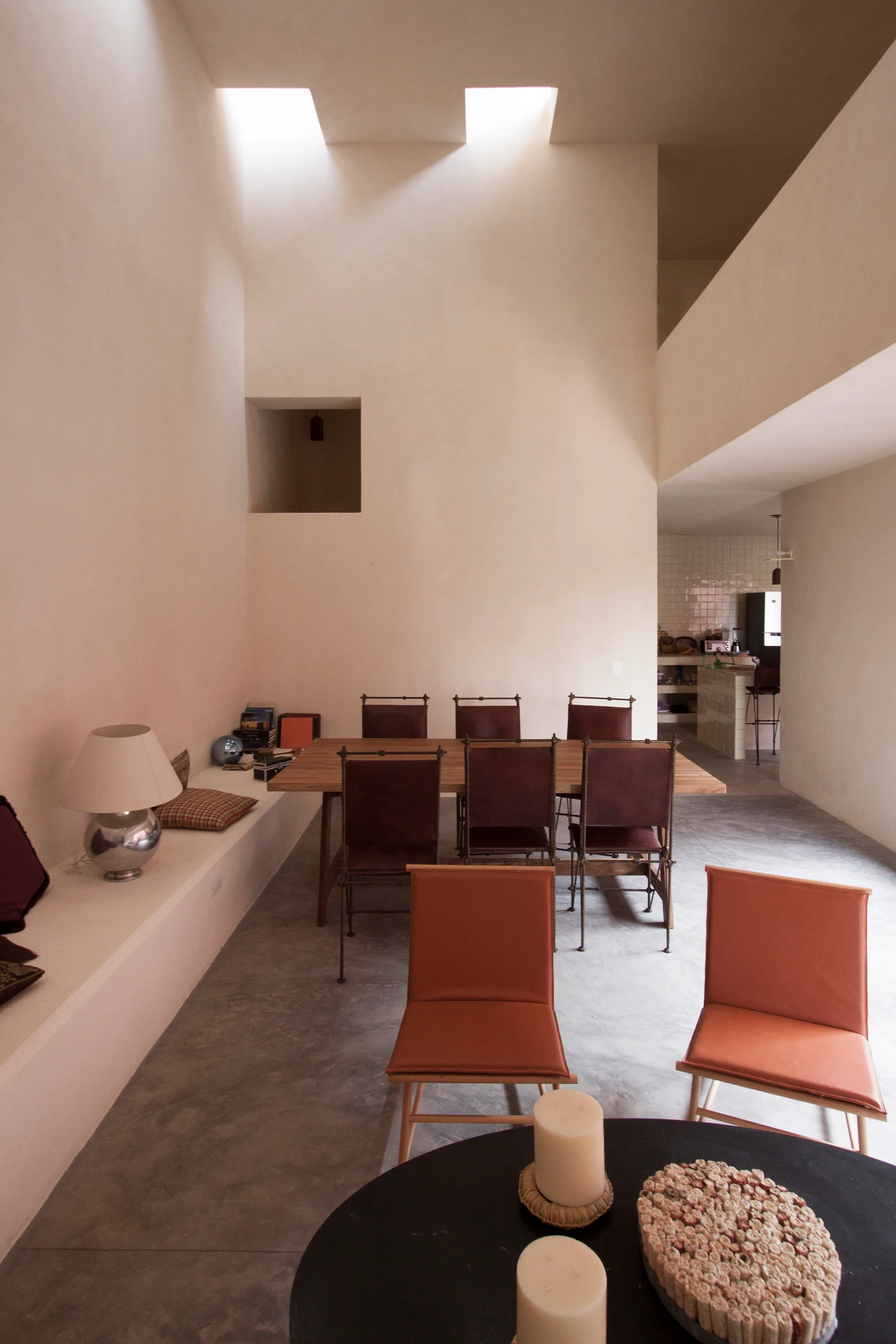 A minimalist dining room features a wooden table and leather chairs under high ceilings with skylights and a long built-in plaster bench.