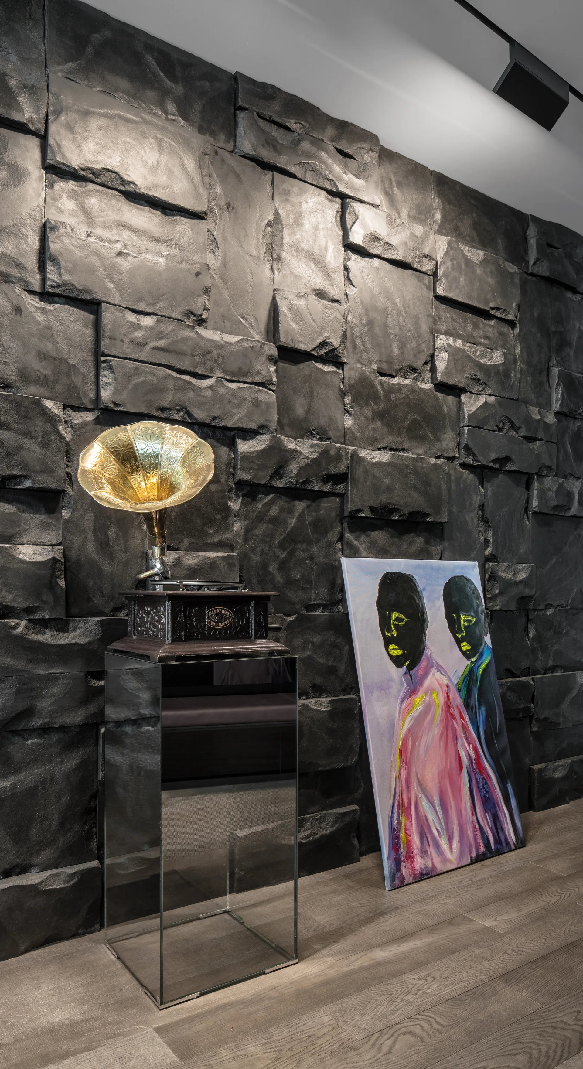 A vintage gramophone on a mirrored pedestal stands beside a colorful canvas leaning against a rough, dark charcoal stone wall.