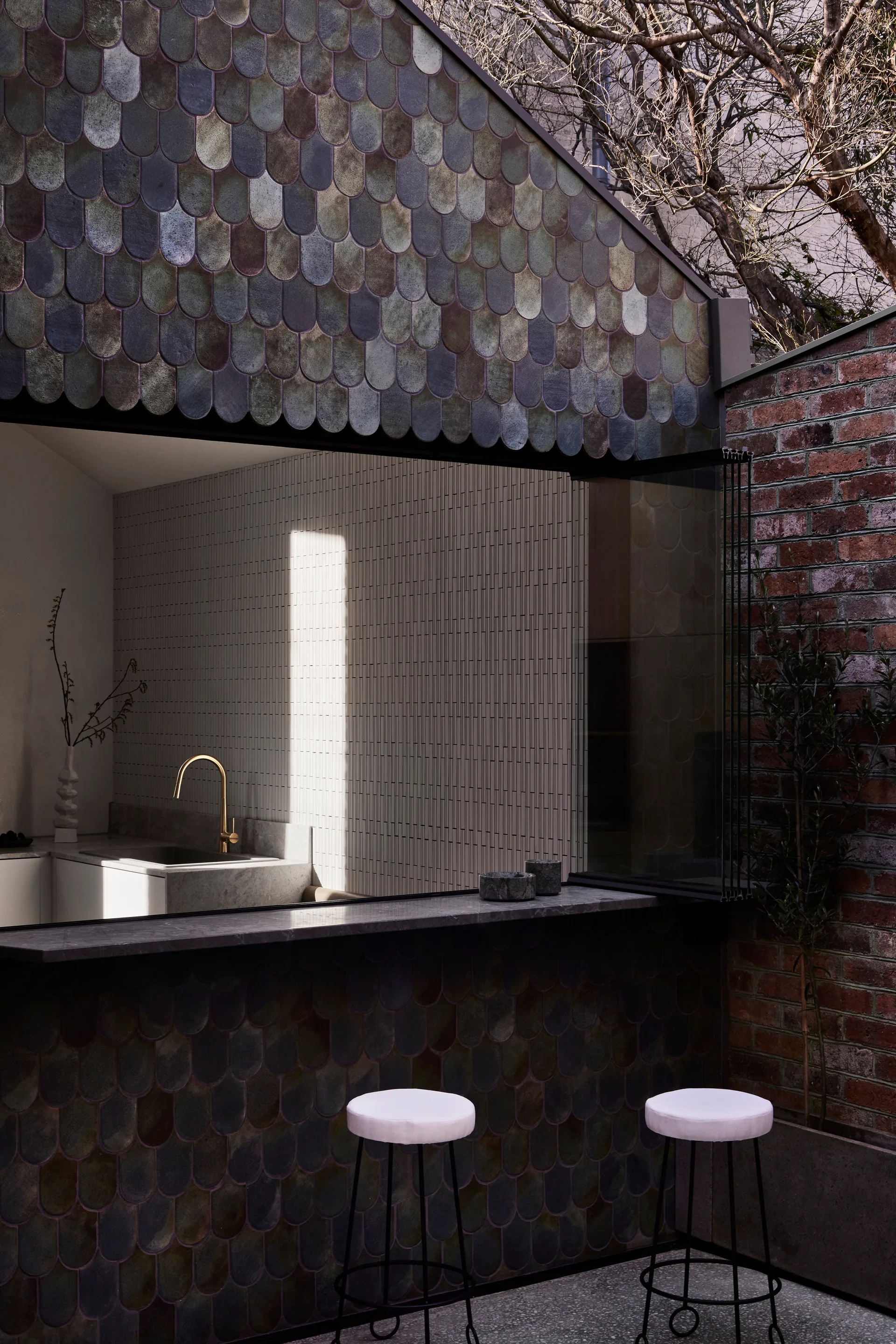 A modern patio features a pass-through window to a kitchen surrounded by iridescent green scalloped tiles, a brick wall, and two bar stools.