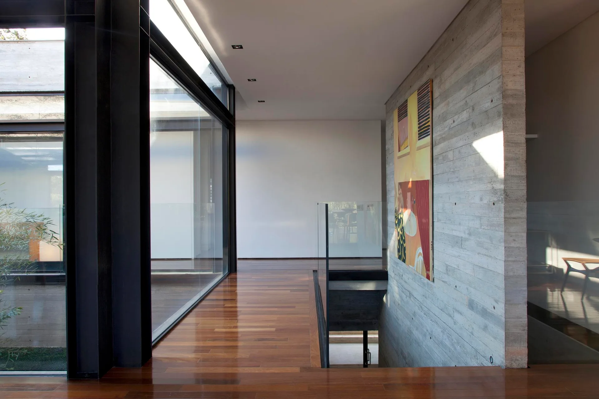 A modern hallway landing features polished wood flooring, a textured board-formed concrete wall with artwork, and a glass-enclosed staircase.
