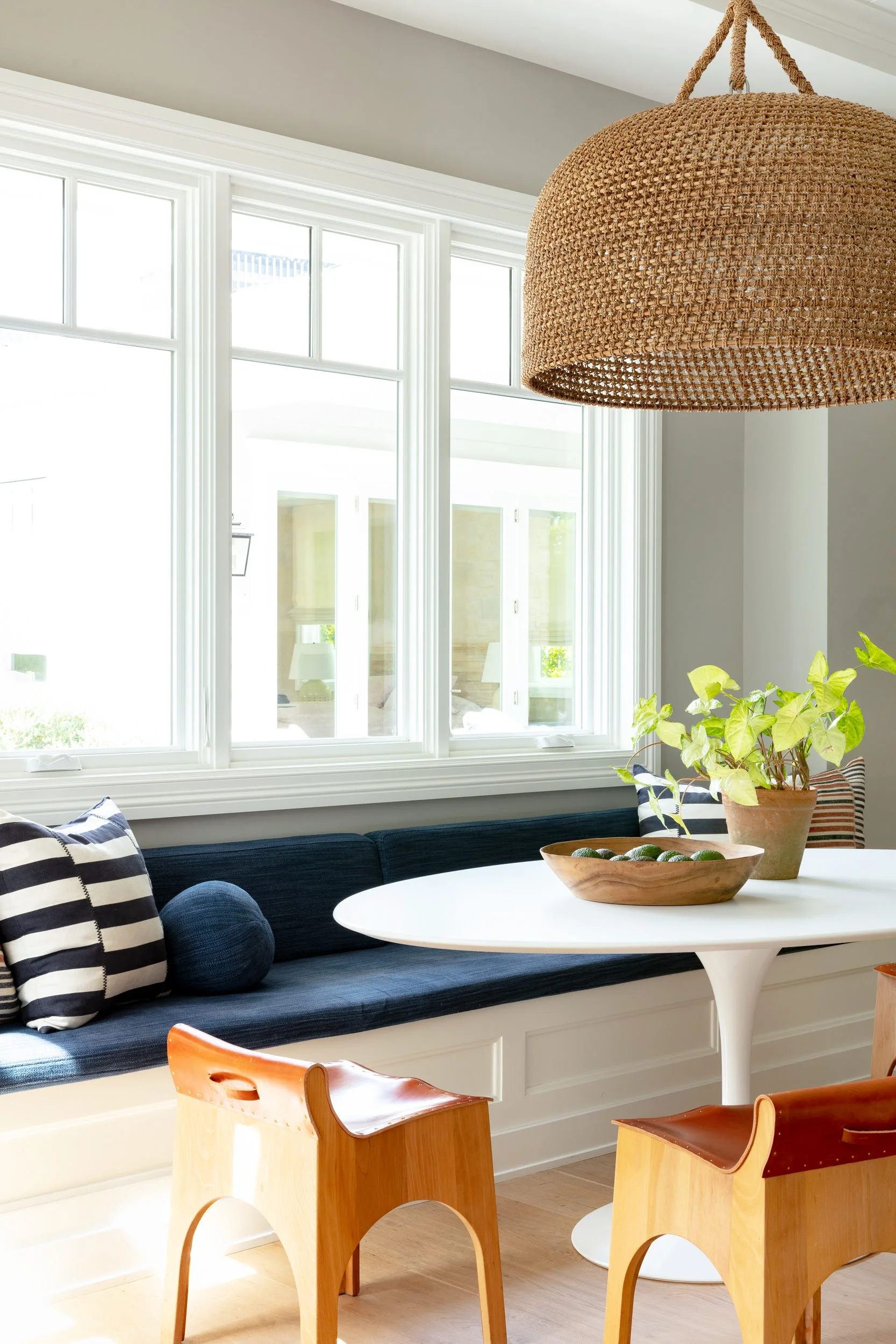 A bright dining nook features a built-in bench with blue cushions, a white oval table, leather chairs, and a large woven pendant light.