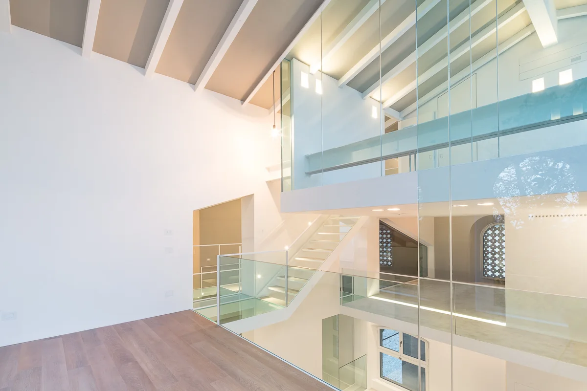 A multi-level modern interior features a white floating staircase with glass railings, exposed ceiling beams, and warm wood flooring.