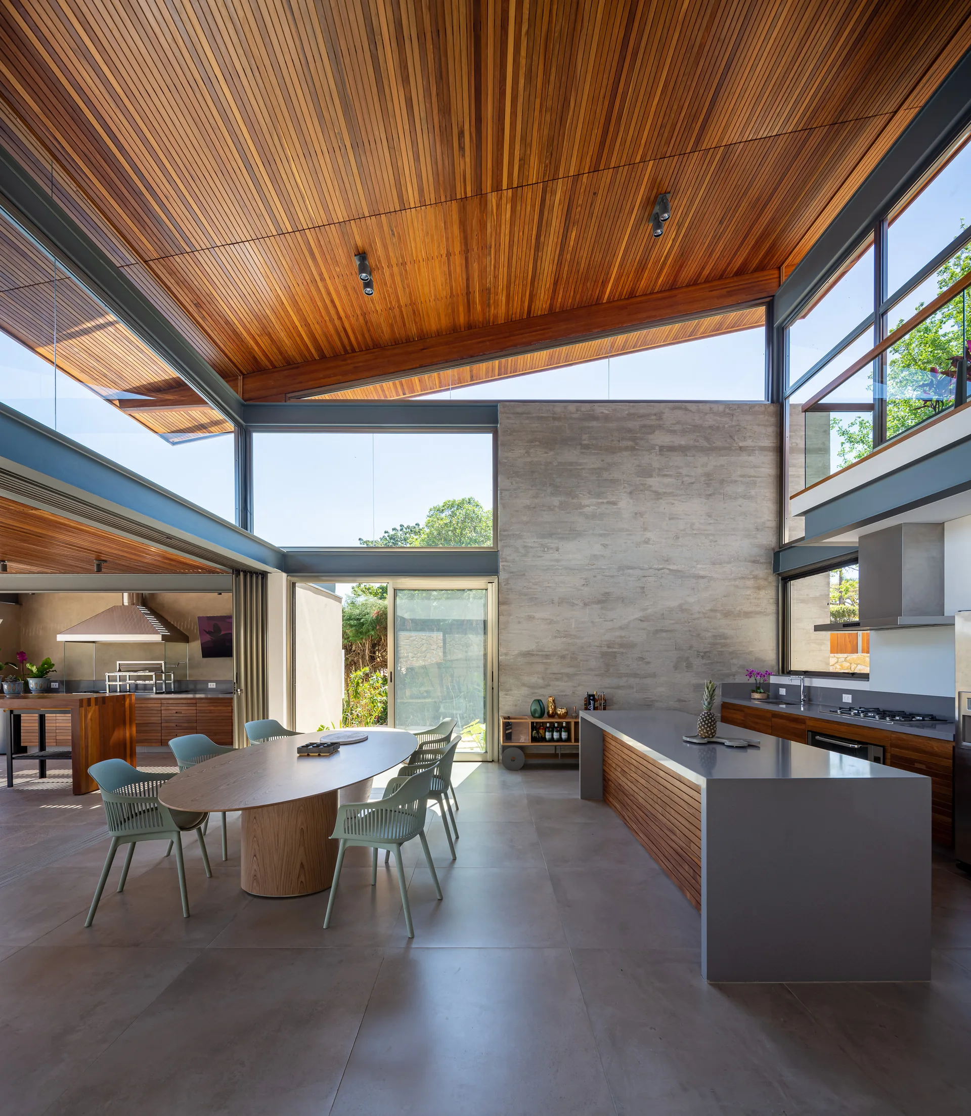 A modern open-concept kitchen features a slanted wood-slat ceiling, an exposed concrete wall, a large grey island, and an oval dining table.