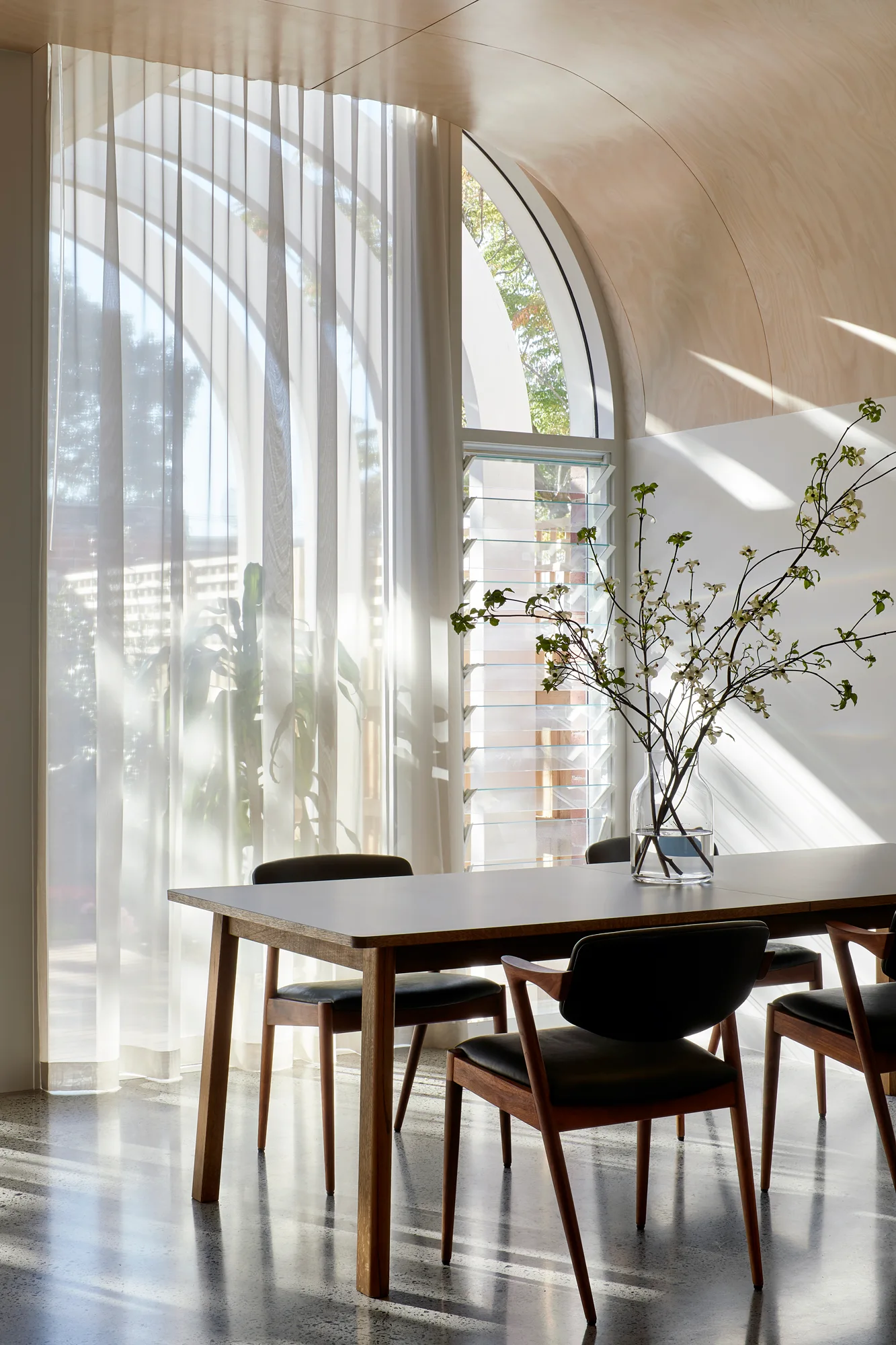 A modern dining room features a wooden table and chairs beneath a curved wood paneled ceiling and a large arched window with sheer white curtains.