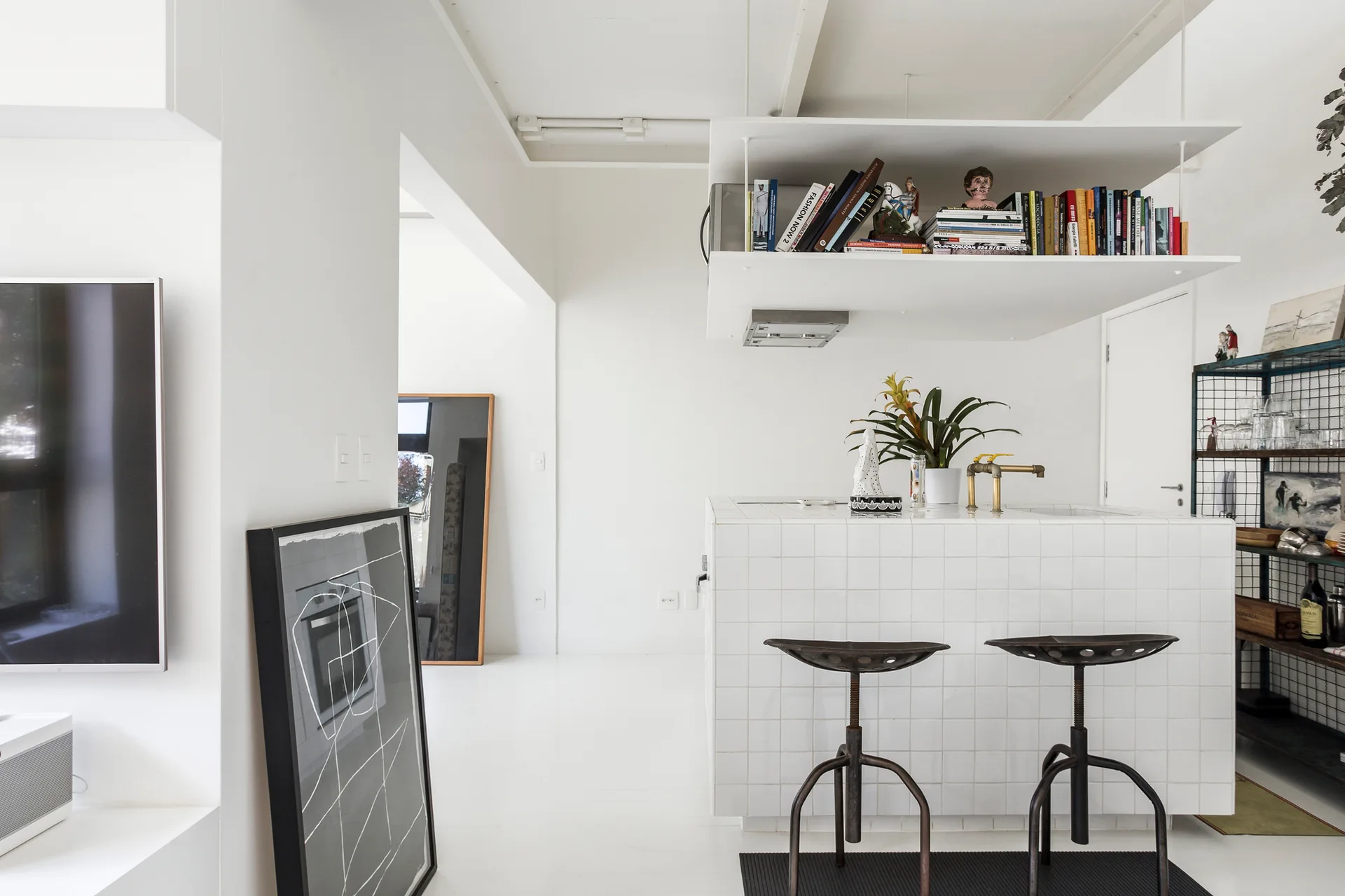 A minimalist white kitchen featuring a square-tiled island with vintage metal bar stools and an overhead suspended shelf filled with books.