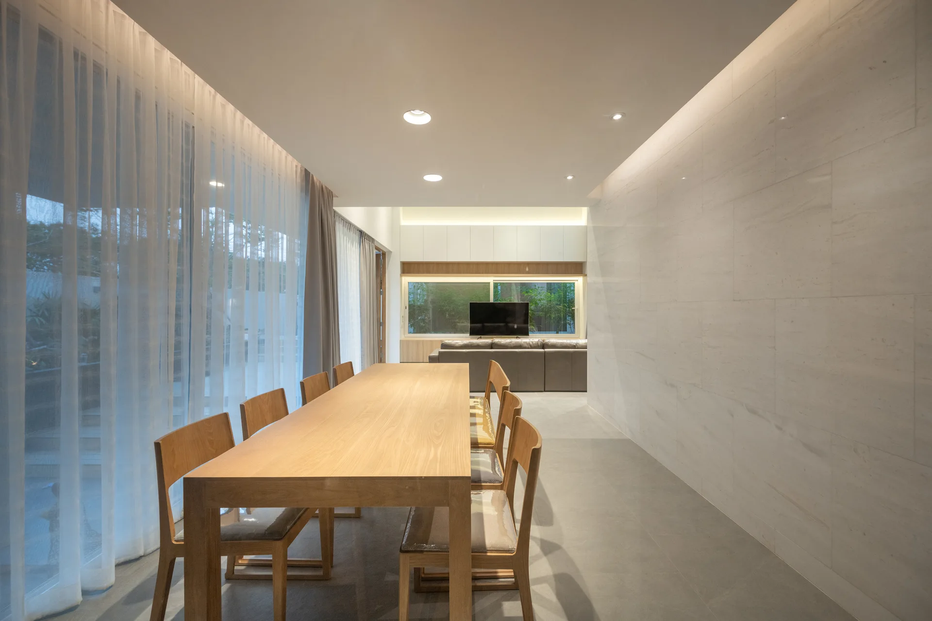 A long natural wood dining table with chairs stands in a minimalist room beside a grey stone-tiled wall and sheer white curtains.