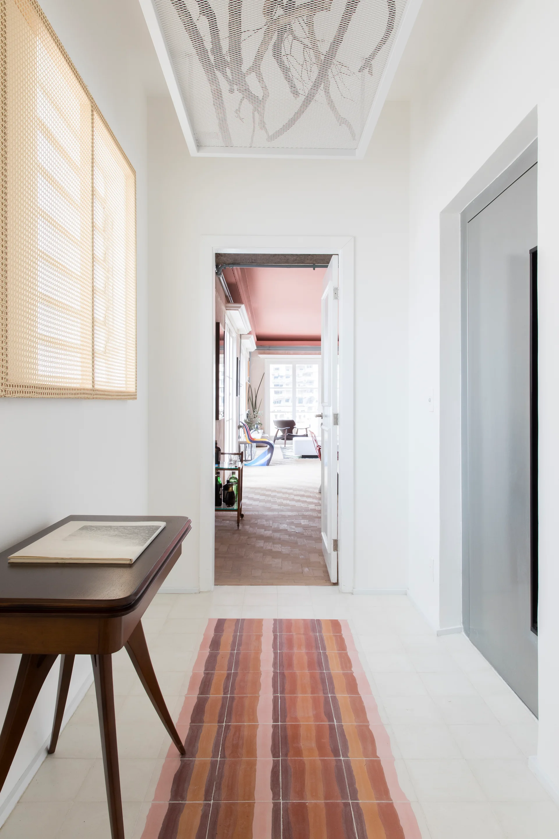 A bright white hallway features a dark wood console table, a cane wall screen, a patterned mesh ceiling panel, and a terracotta-striped floor runner.