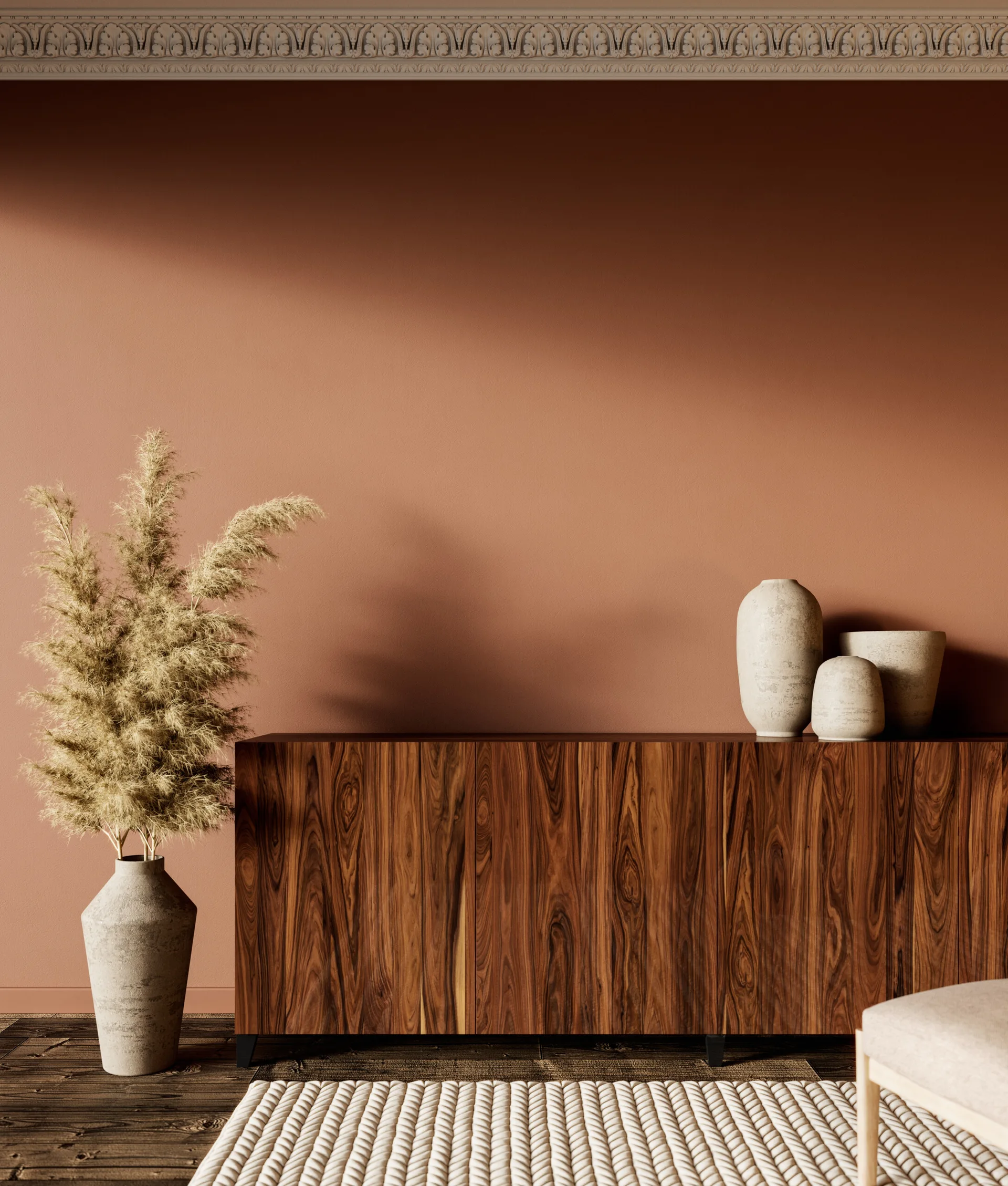 A polished wood grain cabinet sits against a terracotta wall with white crown molding, flanked by a tall vase of dried pampas grass.