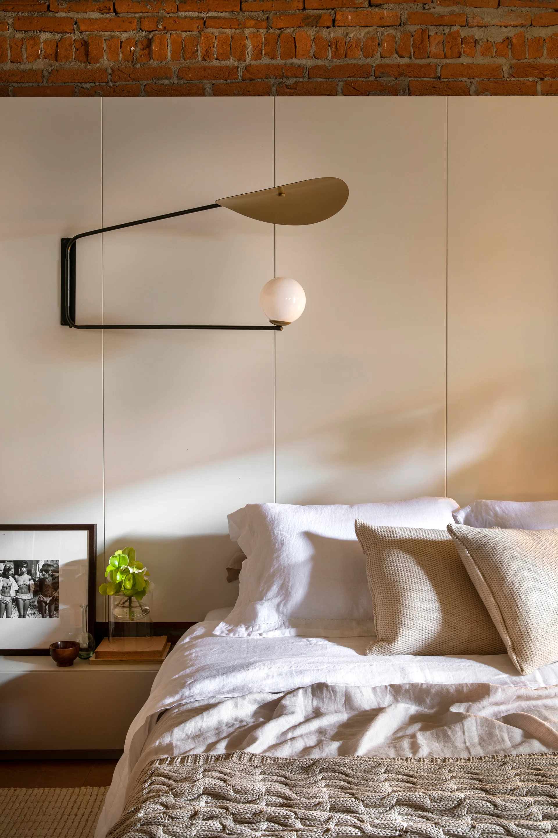 A bed with white linen sheets and knit pillows sits below a light wood paneled wall topped with exposed brick and a modern black wall sconce.