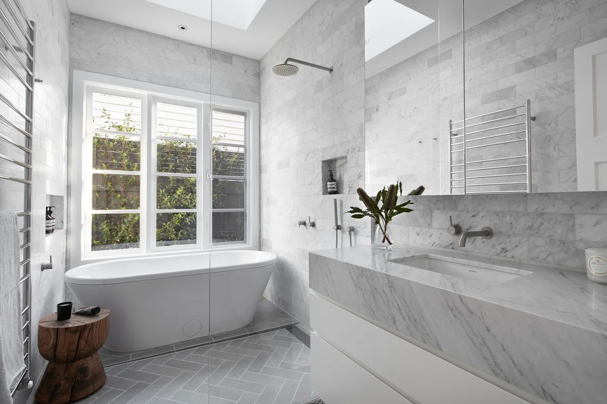 A modern bathroom featuring a white freestanding bathtub by a large window, a marble vanity, and grey herringbone floor tiles.
