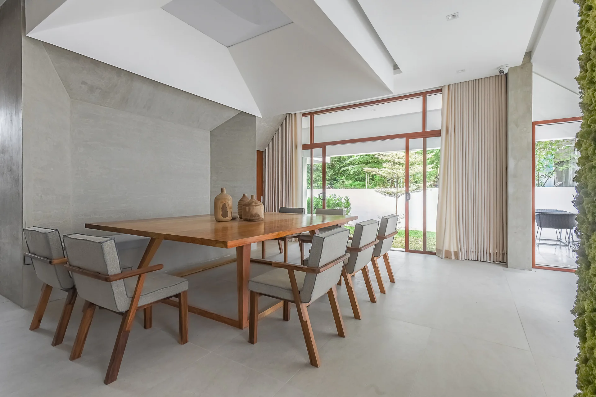 A modern dining room featuring a large wooden table with six upholstered chairs, grey concrete walls, and sliding glass doors that open to a garden.