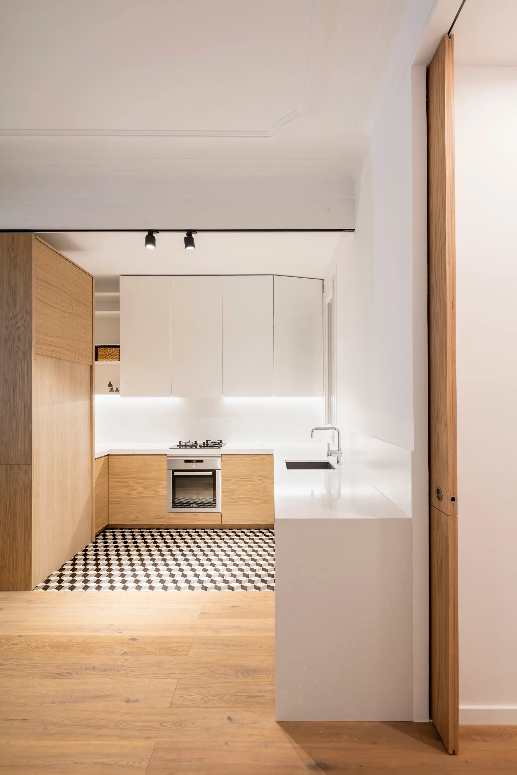 A minimalist kitchen features geometric patterned floor tiles, light wood and white cabinetry, a built-in oven, and a white countertop.