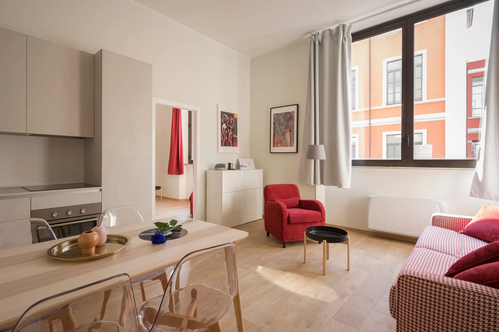 A bright apartment living and dining area featuring a light wood table with clear plastic chairs, a red armchair, and a red patterned sofa.