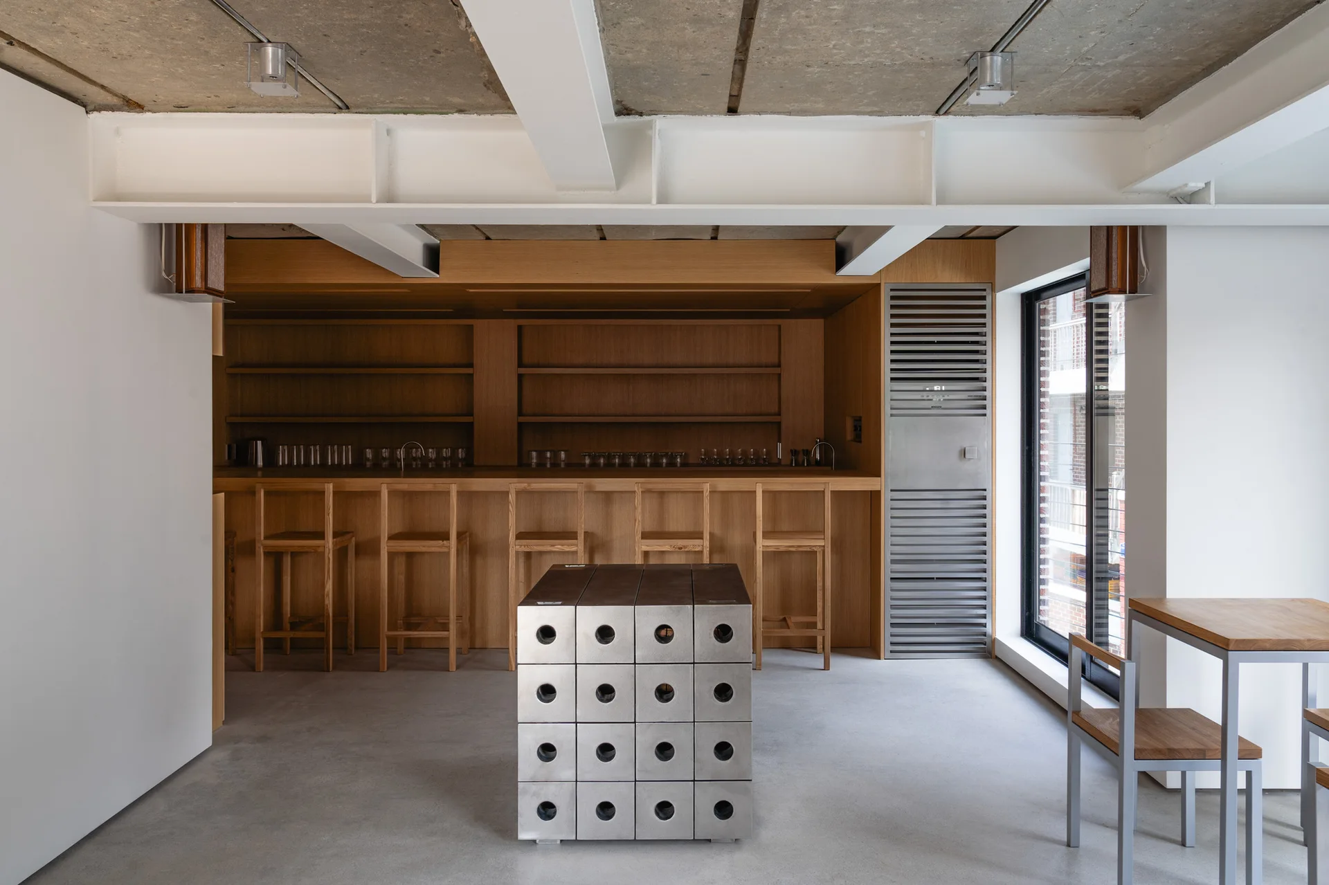 A minimalist commercial bar space featuring a long wooden counter with stools, a geometric metal center block, and an exposed concrete ceiling.