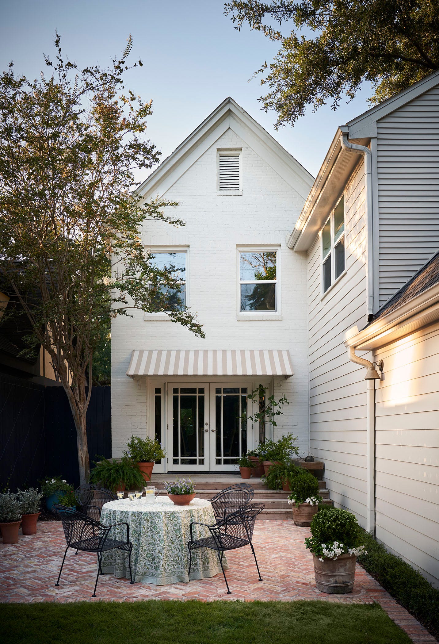 A red herringbone brick patio features a round dining table with a patterned tablecloth and black metal chairs outside a white house.